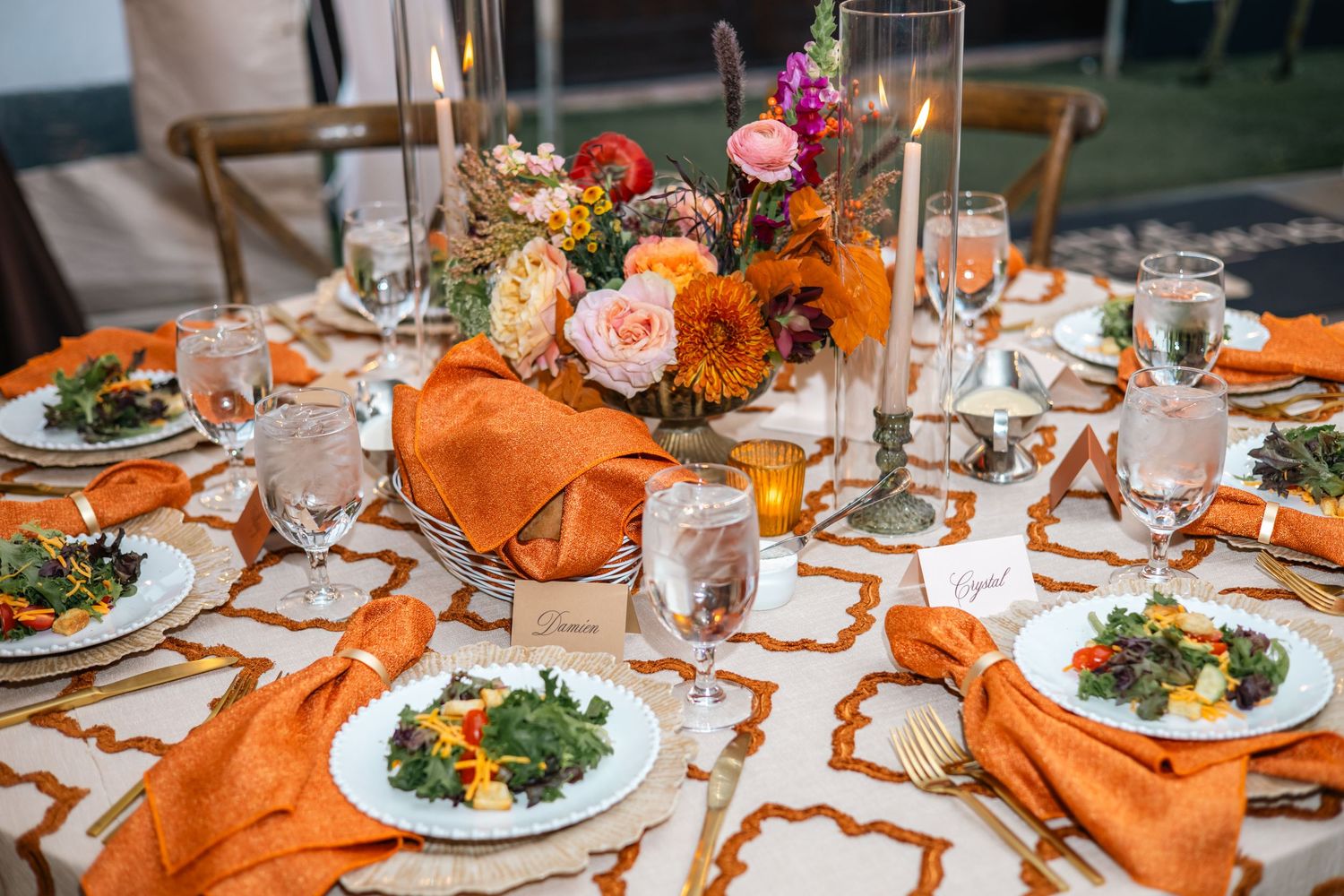 An elegant table setting with orange linens, floral centerpieces, and plated meals at an autumn-themed dinner party.