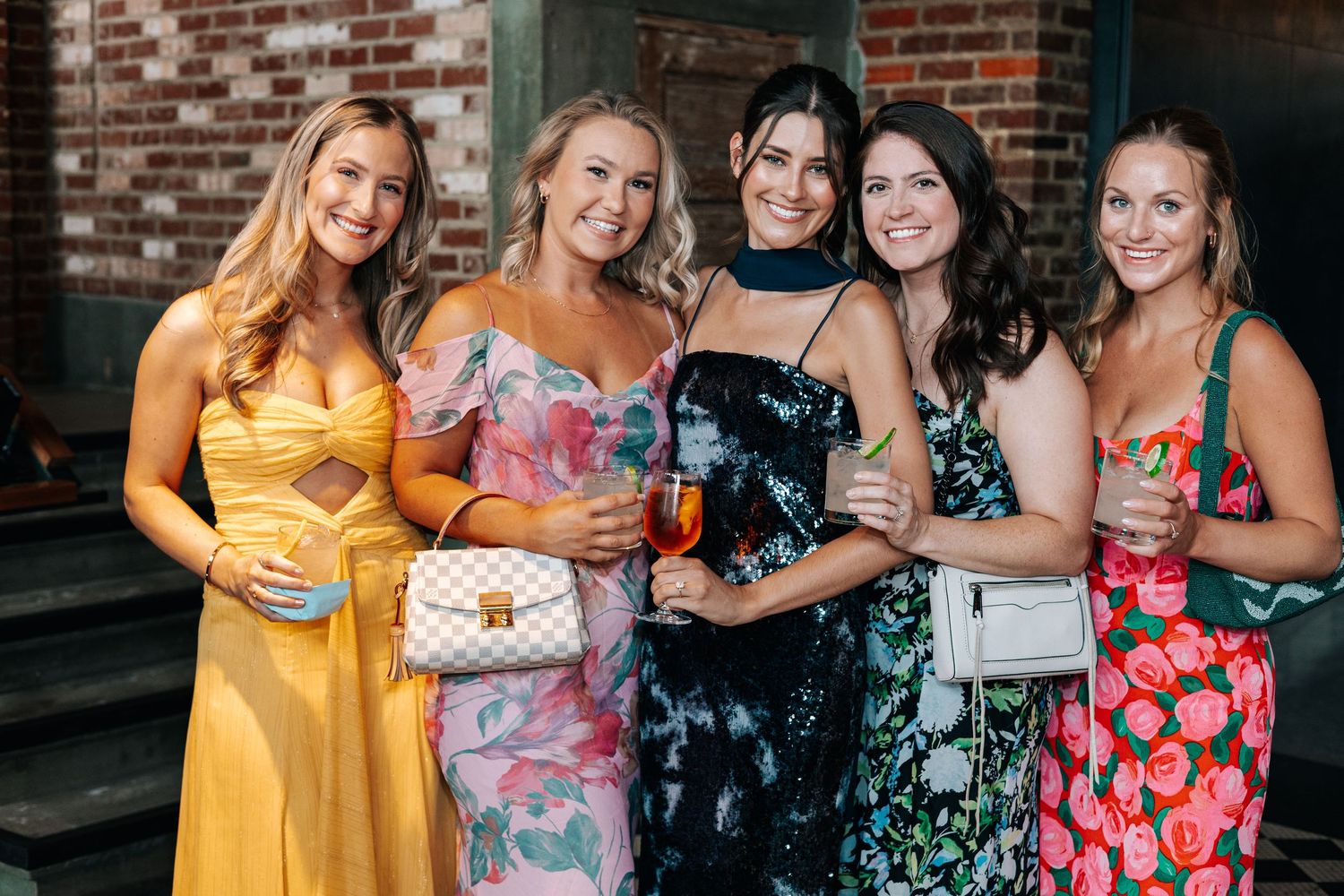 A group of friends in colorful summer dresses pose together at a social gathering against a brick wall backdrop.