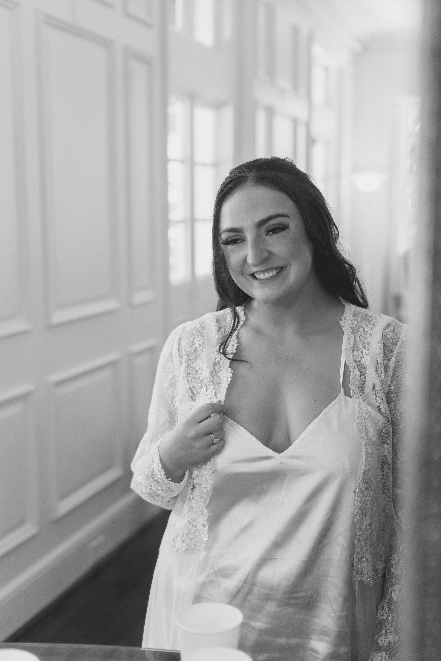 Black and white photo of a smiling bride in a lace robe and satin slip dress during wedding preparations.