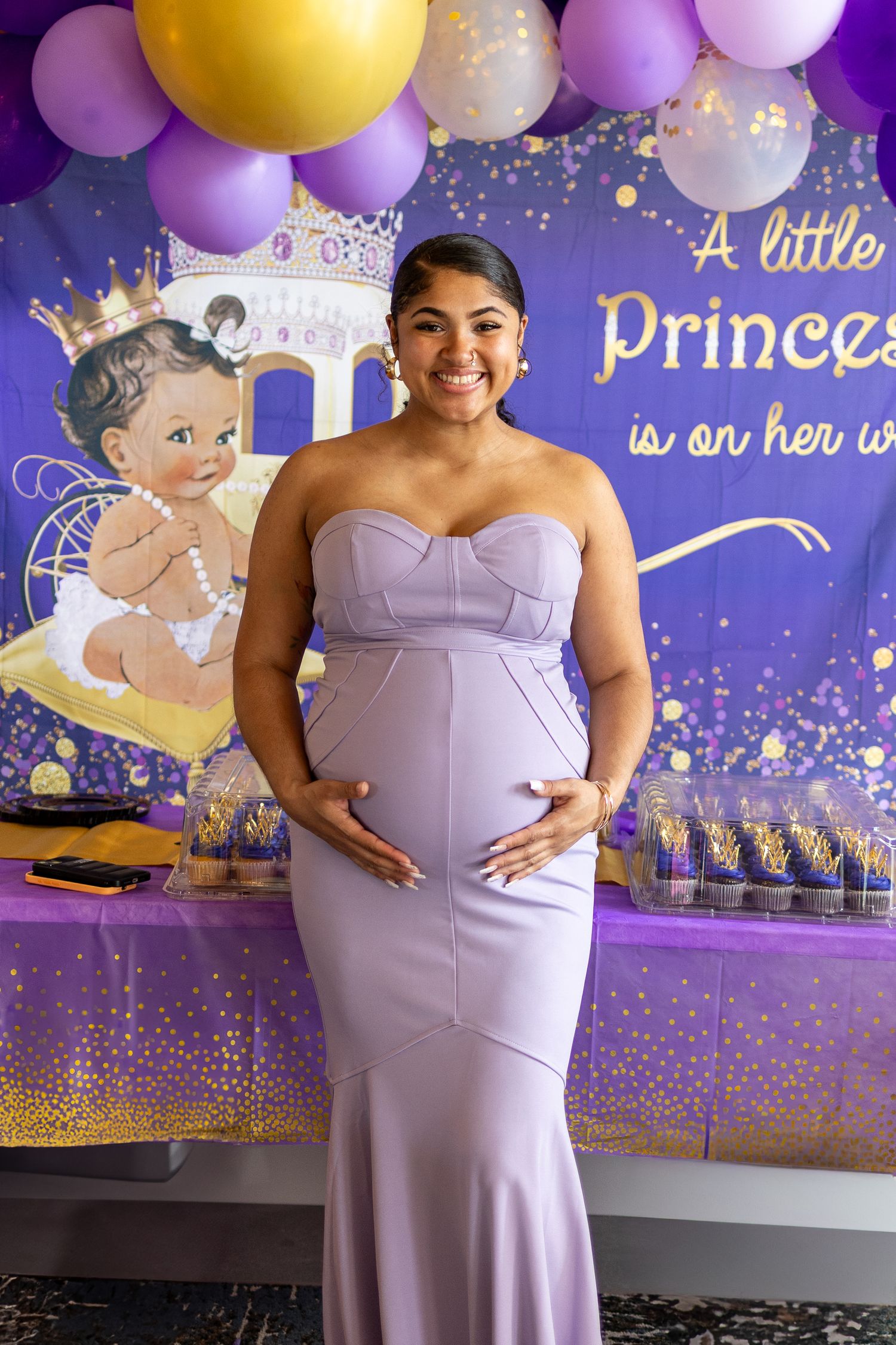A lilac-colored maternity dress shown in front of a purple and gold balloon backdrop with princess-themed decorations.