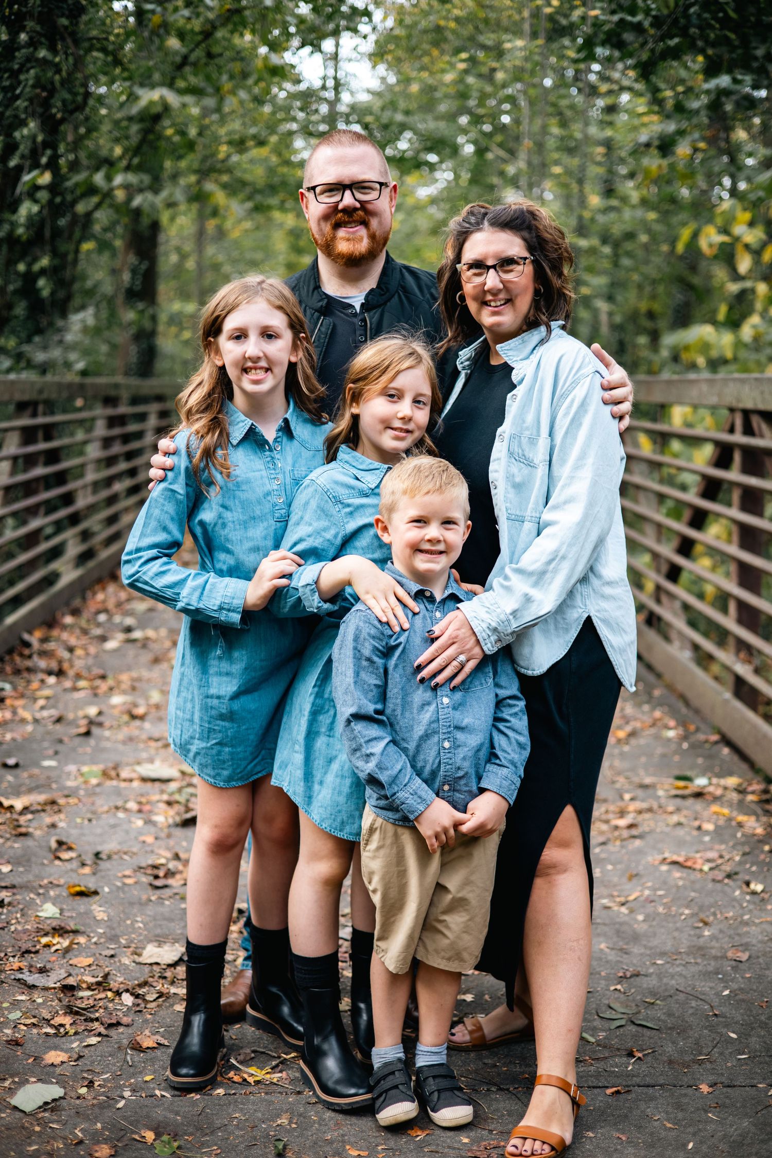 A family of five wearing coordinating blue outfits poses together on a wooden bridge surrounded by autumn trees.