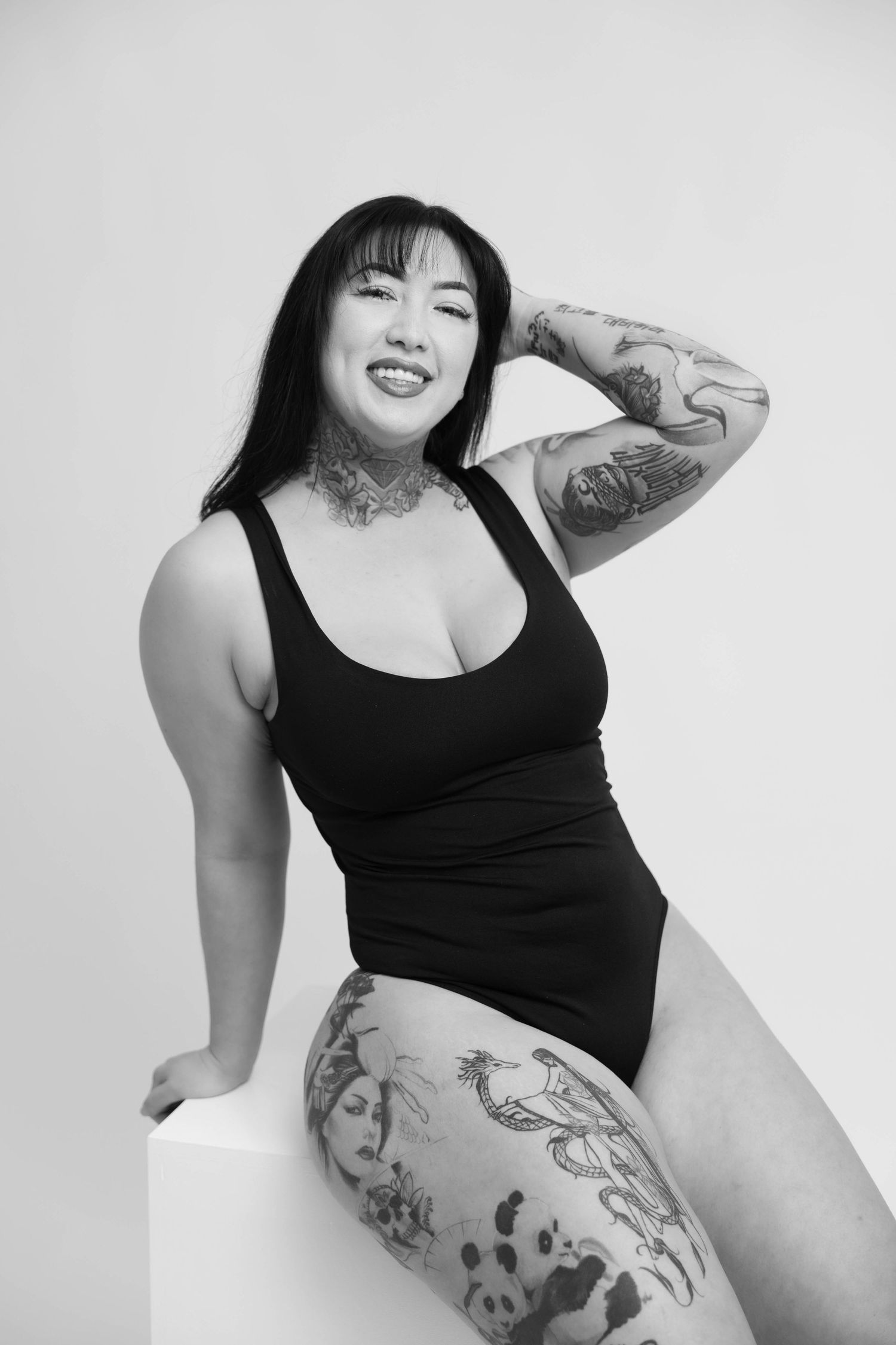 Black and white studio portrait of model in black tank top showing tattoos while posing against white wall.