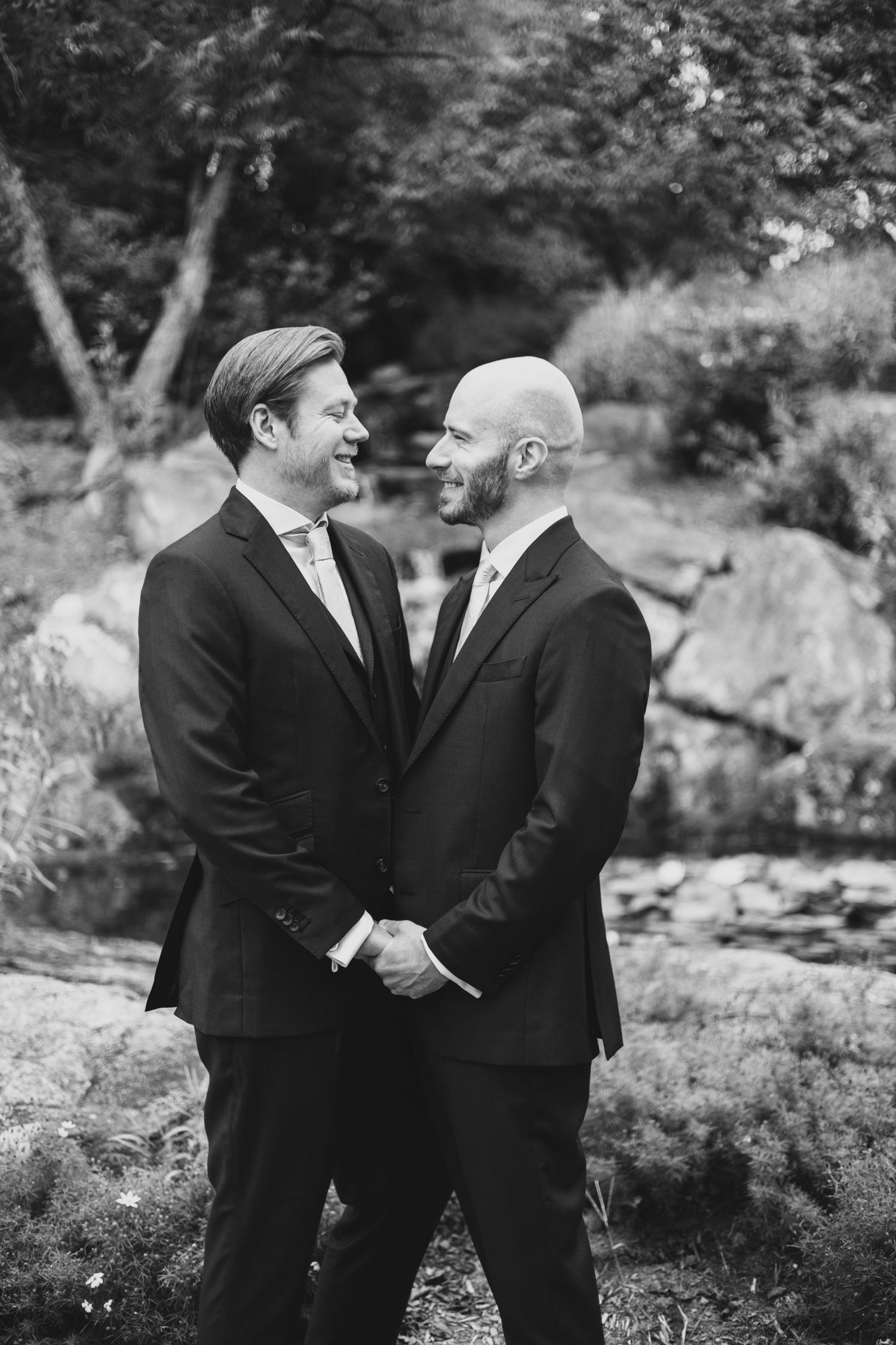 Two people in black suits share joyful laughter together outdoors in a natural setting during a formal occasion.