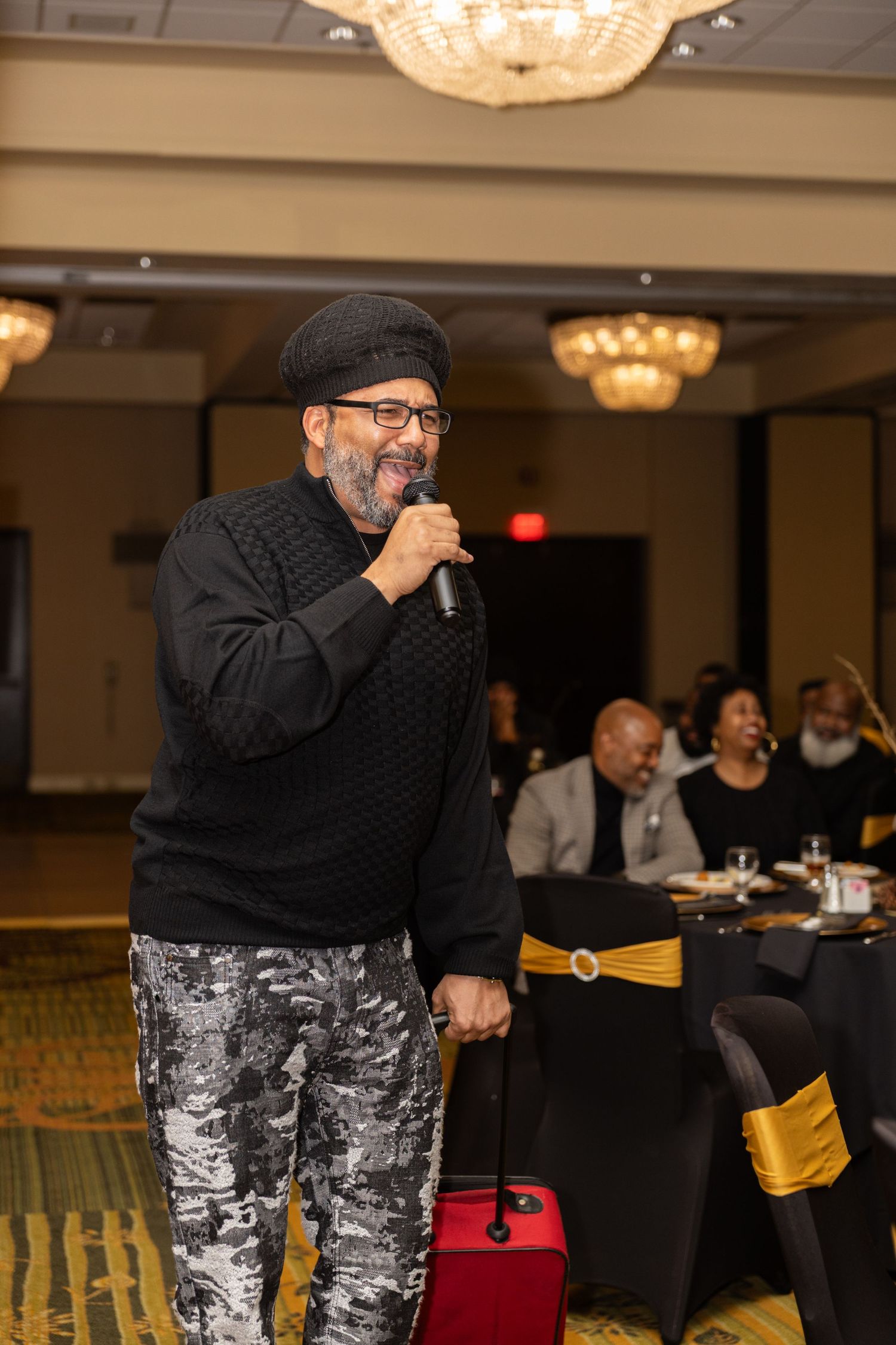A performer in a black top and patterned pants entertains an audience in a banquet hall with crystal chandeliers.