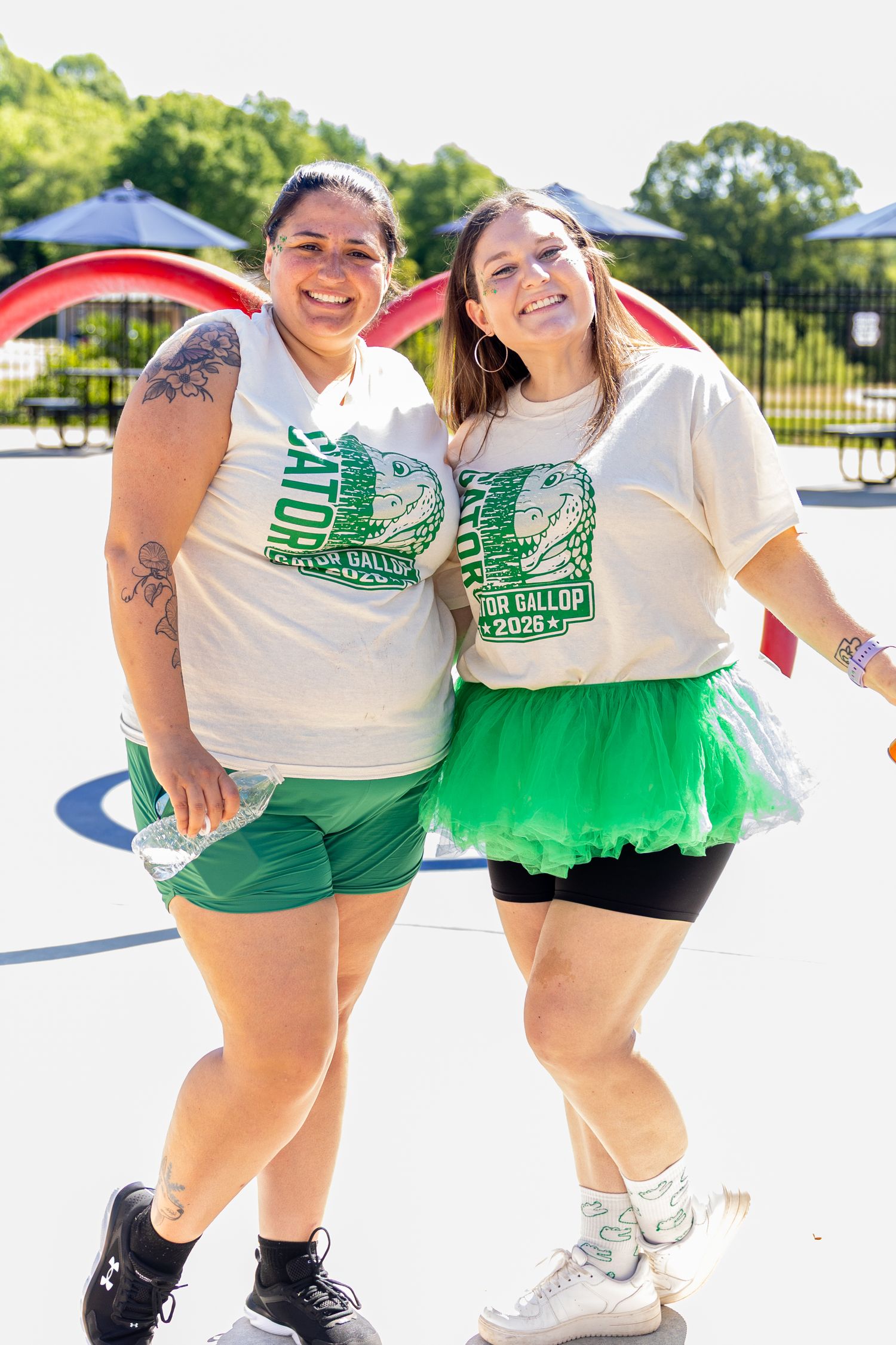 Two women in matching Gator Gallop 2025 shirts smile together on a sunny playground, one wearing a green tutu skirt.