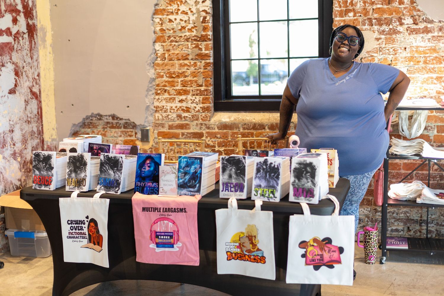 Woman smiling behind a vendor table displaying tote bags and boxed collectible figures at an indoor market event.