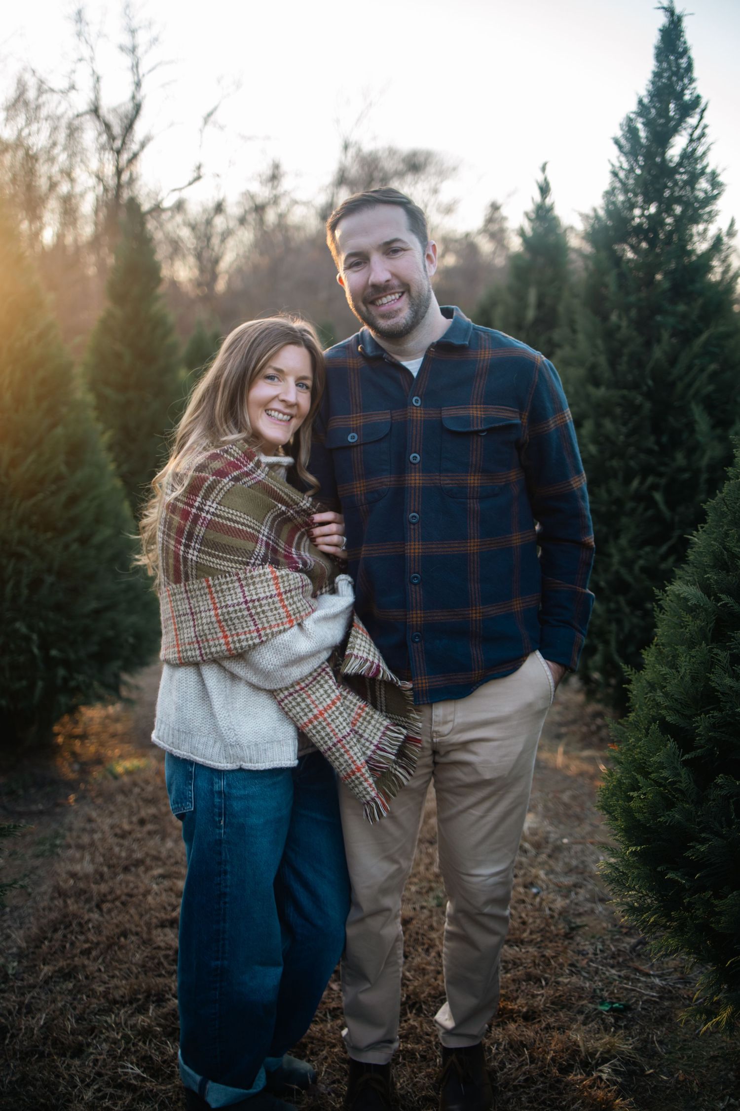 Couple laughing and embracing while surrounded by pine trees during a winter photoshoot.
