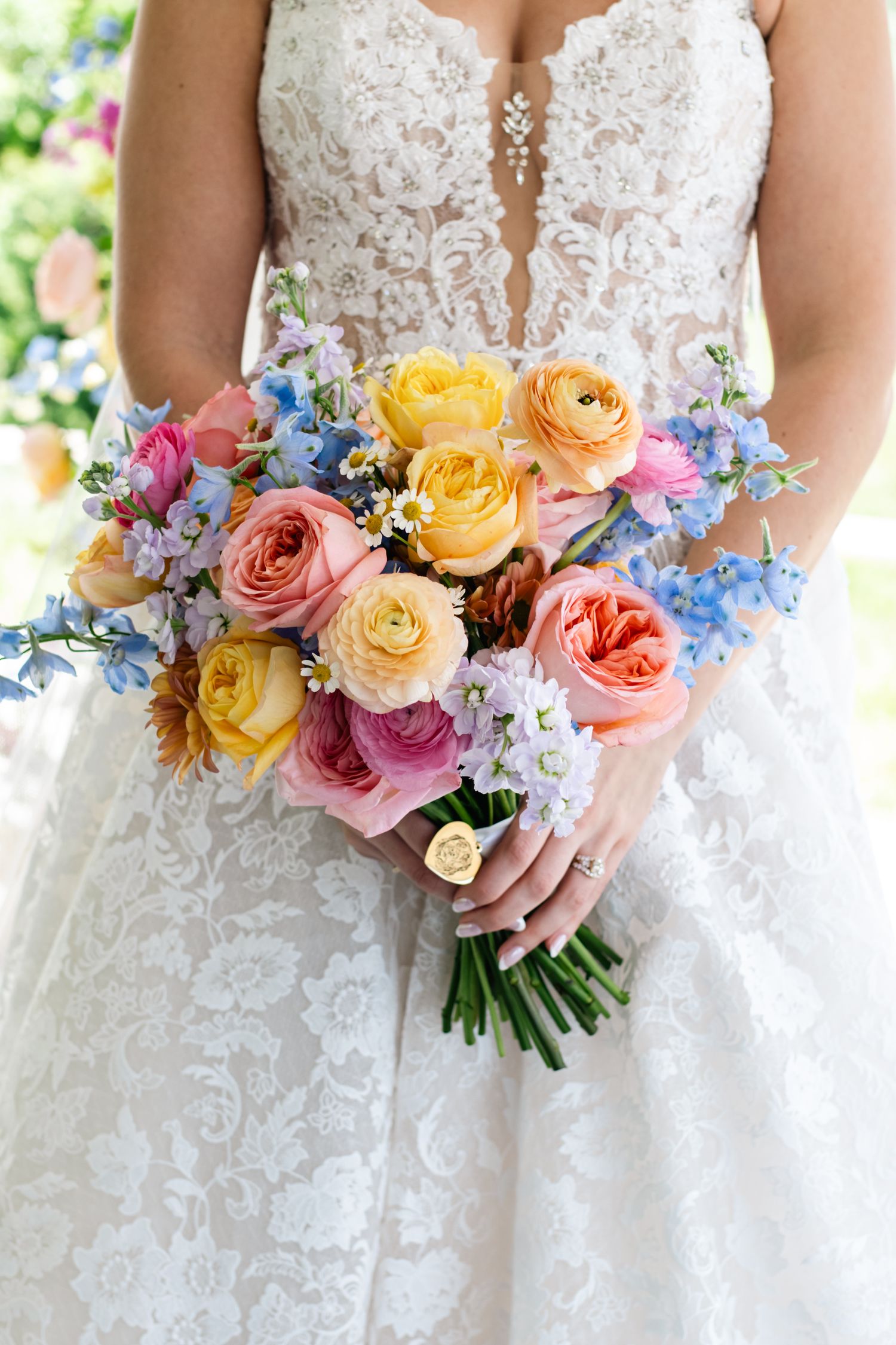 Bride holding colorful wedding bouquet with pink, yellow roses and blue flowers, wearing lace gown.