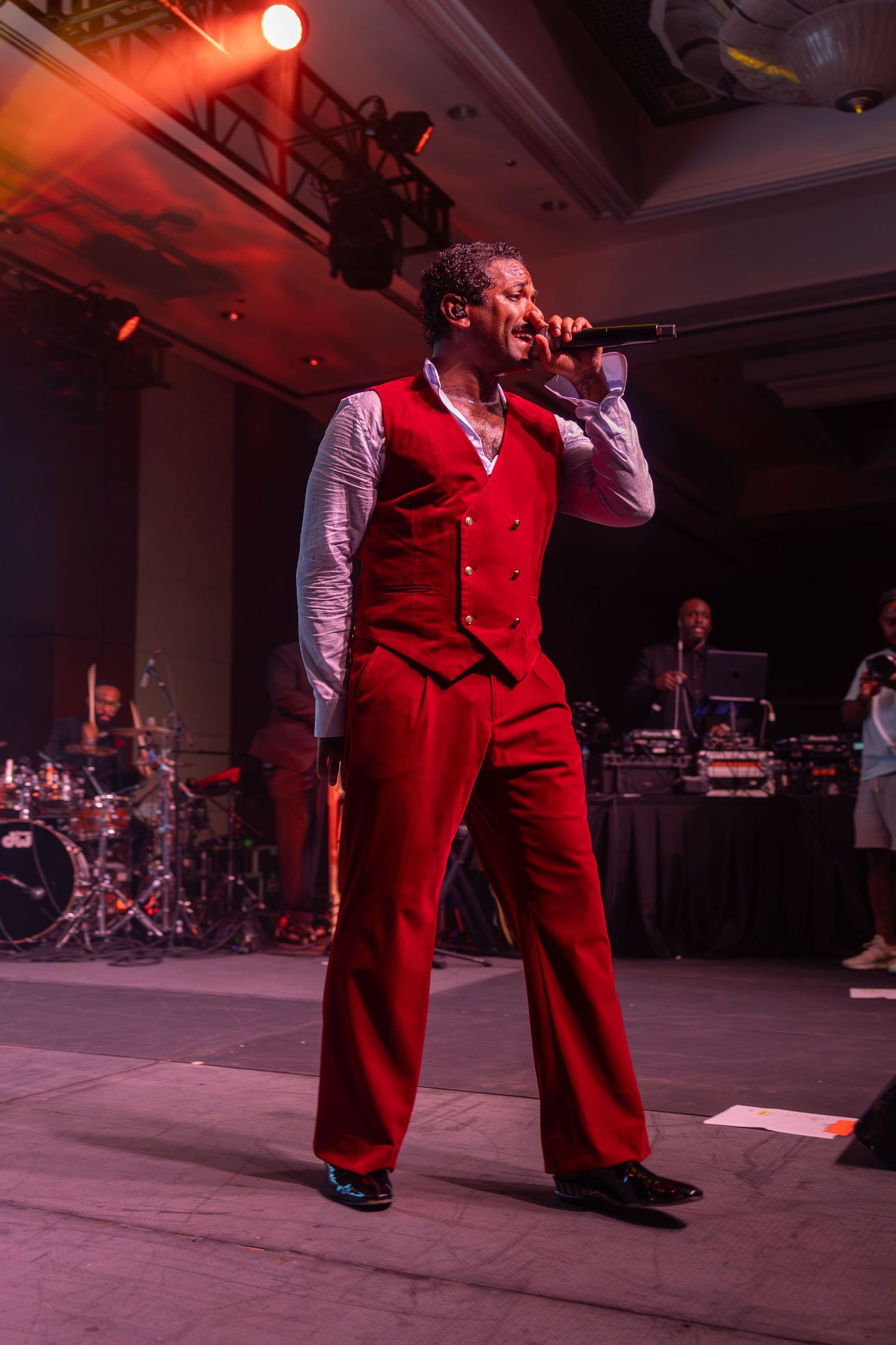 Male singer in red vest and pants performing on stage with microphone under dramatic lighting.