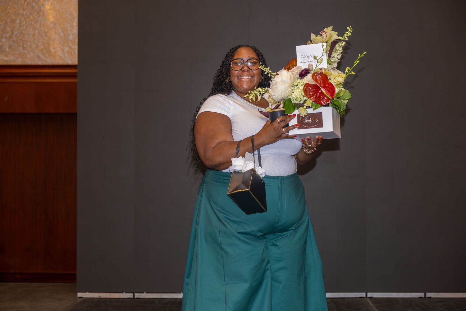 Woman smiling holding flowers and an award at a recognition ceremony in a teal skirt and white top.