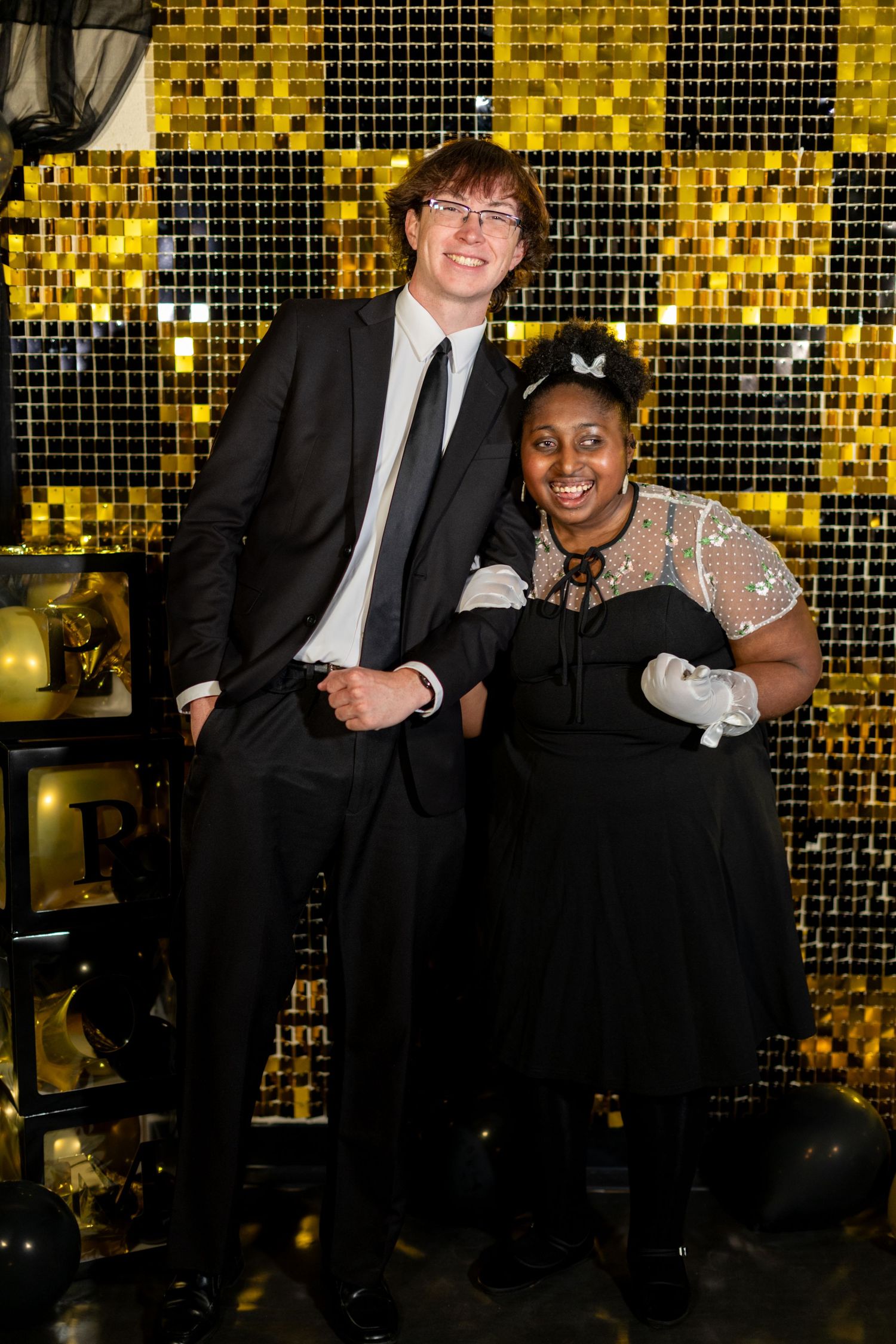Two formally dressed individuals posing together in front of a gold and black sequin backdrop at an event.