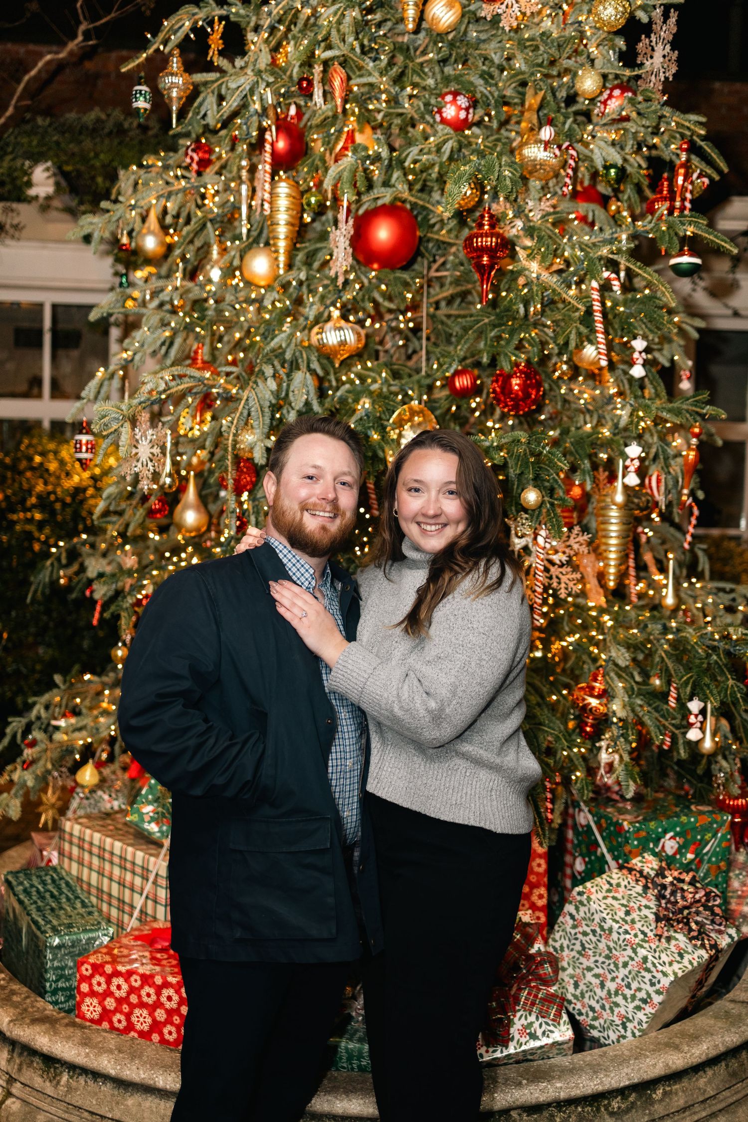 A couple poses together in front of a beautifully decorated Christmas tree with red and gold ornaments.