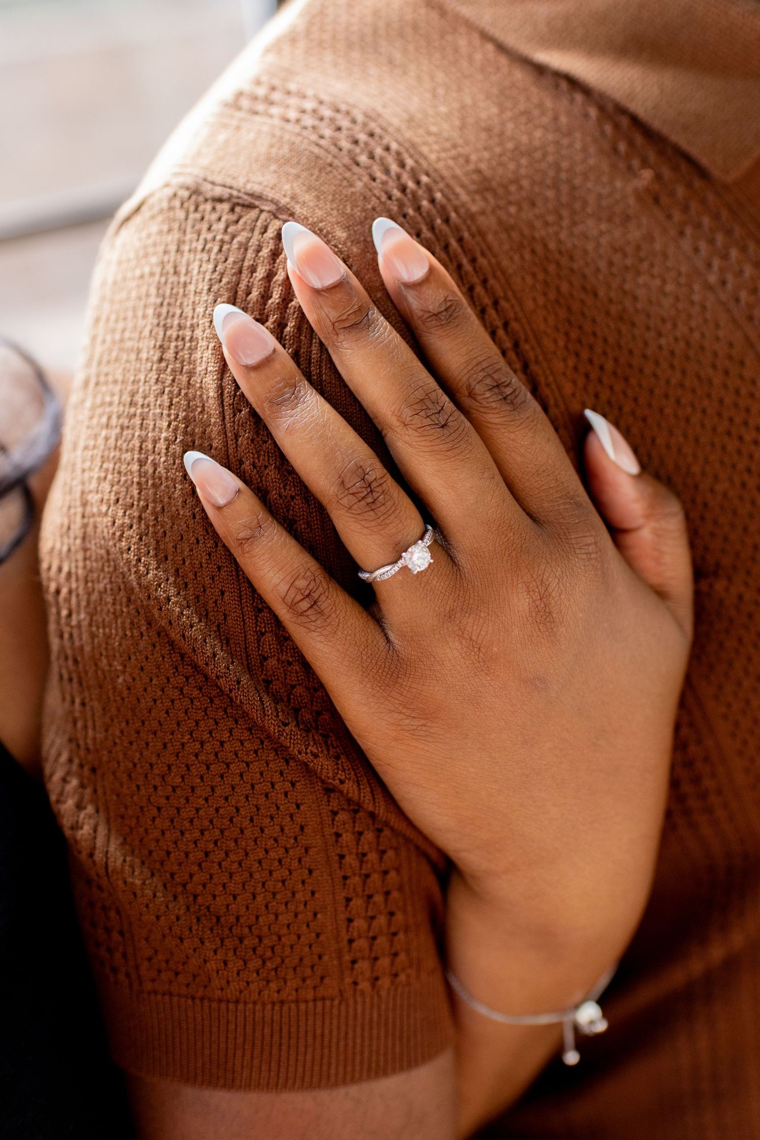 Close-up of a hand with almond nails wearing a diamond engagement ring, resting on a brown knit sweater.