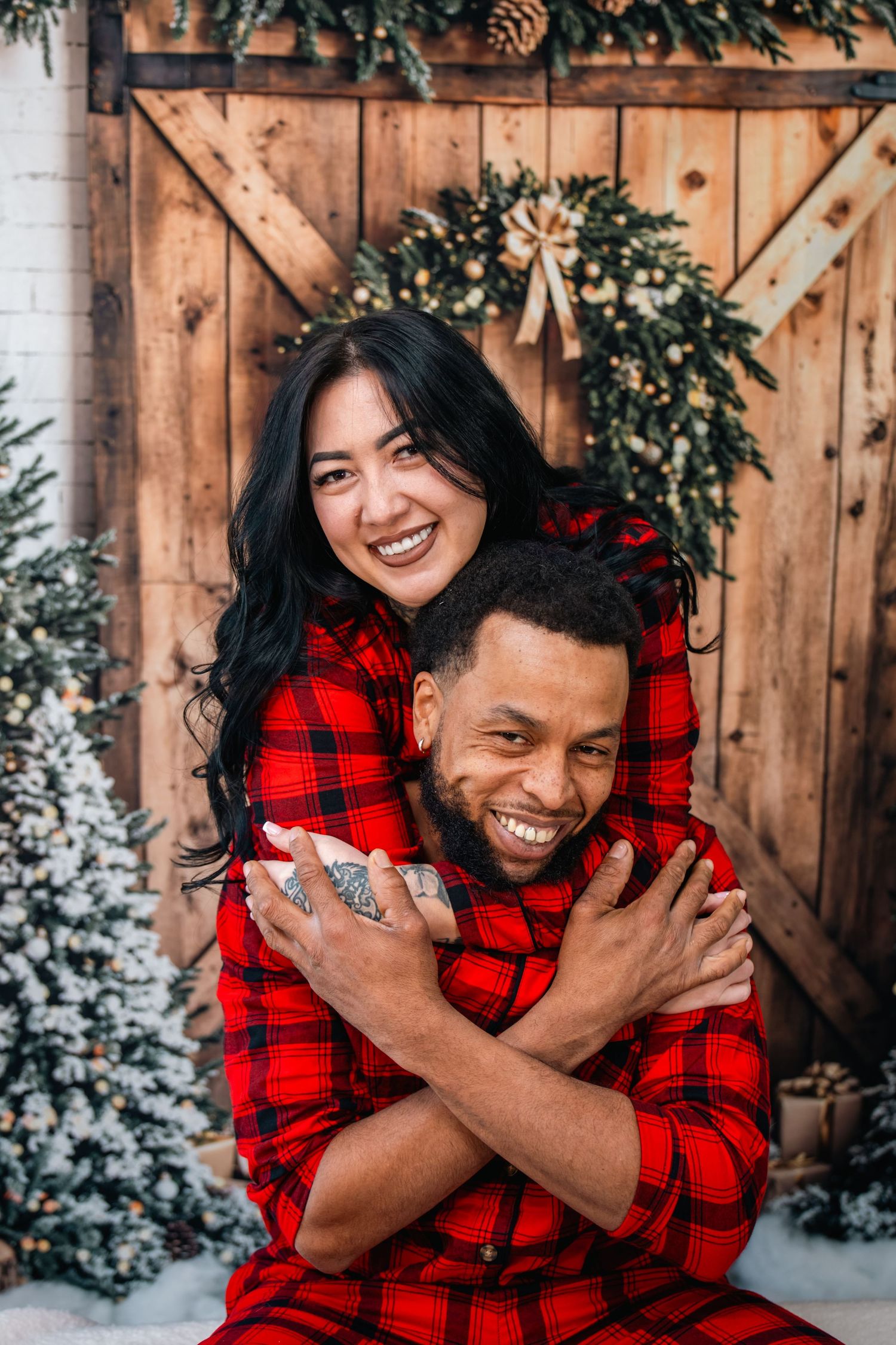 A couple wearing matching red buffalo plaid outfits share joyful laughter in a winter photoshoot against a wooden backdrop.