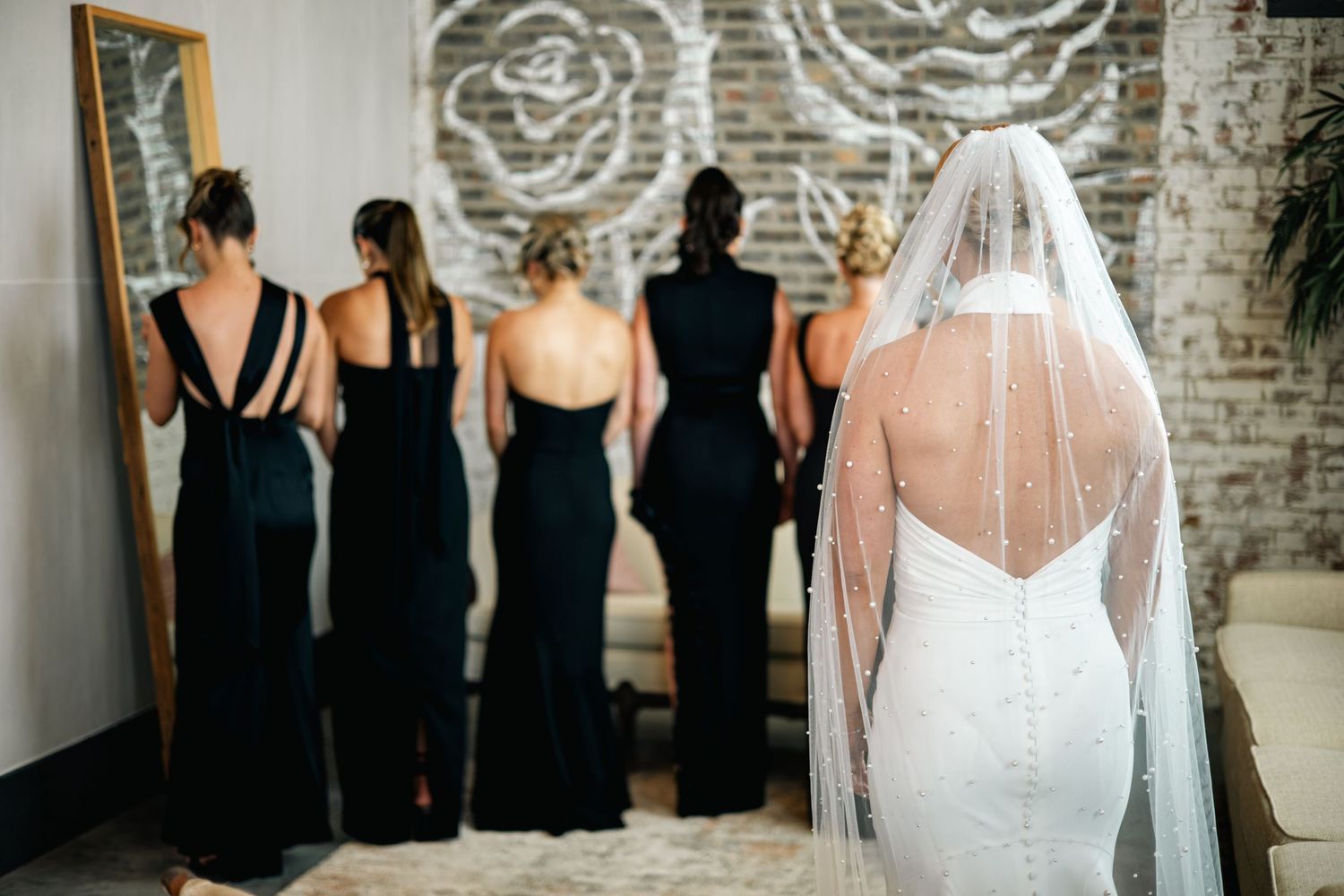 A bridal party poses in black dresses while the bride wears a white gown with veil against a rustic brick wall.