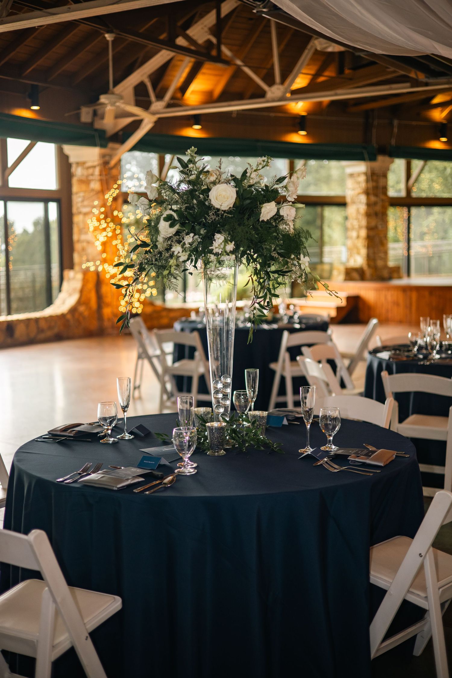Wedding reception venue with black tablecloths and white chairs under rustic wooden beams and soft lighting.