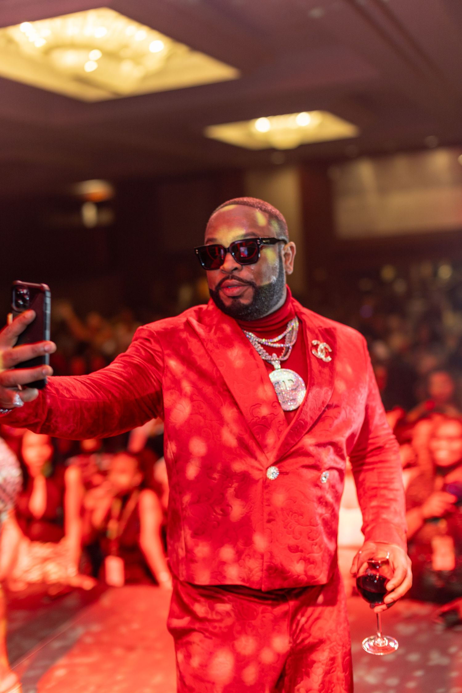 Man in red velvet suit taking selfie while holding wine glass at upscale party event.