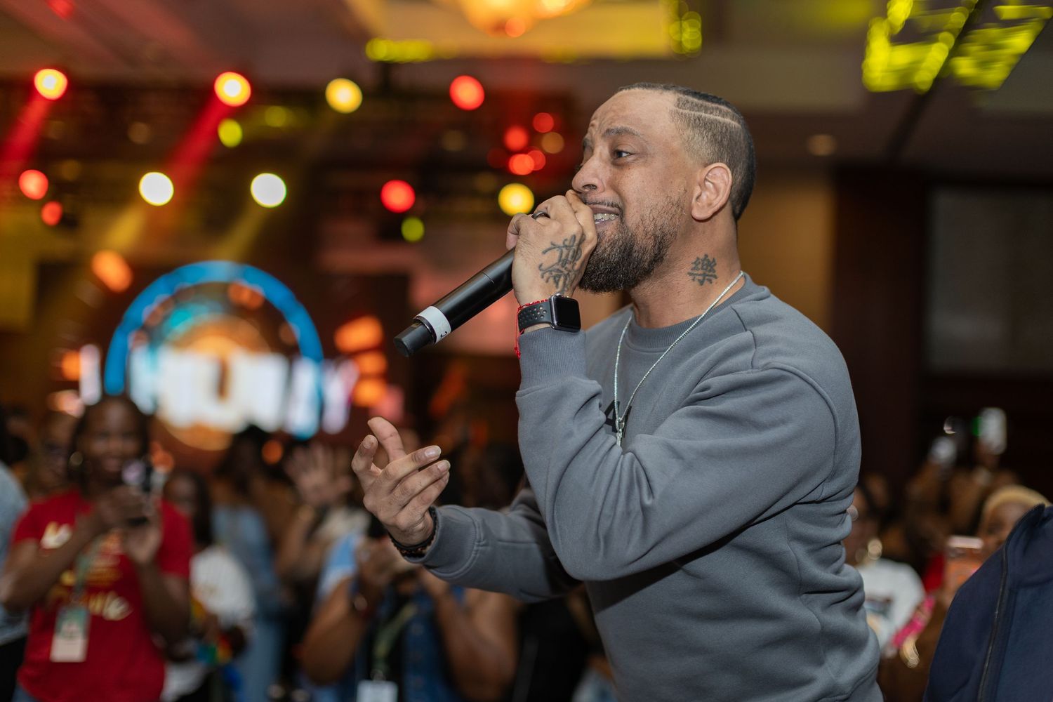 Tattooed male performer singing into microphone at lively indoor event with crowd in background.