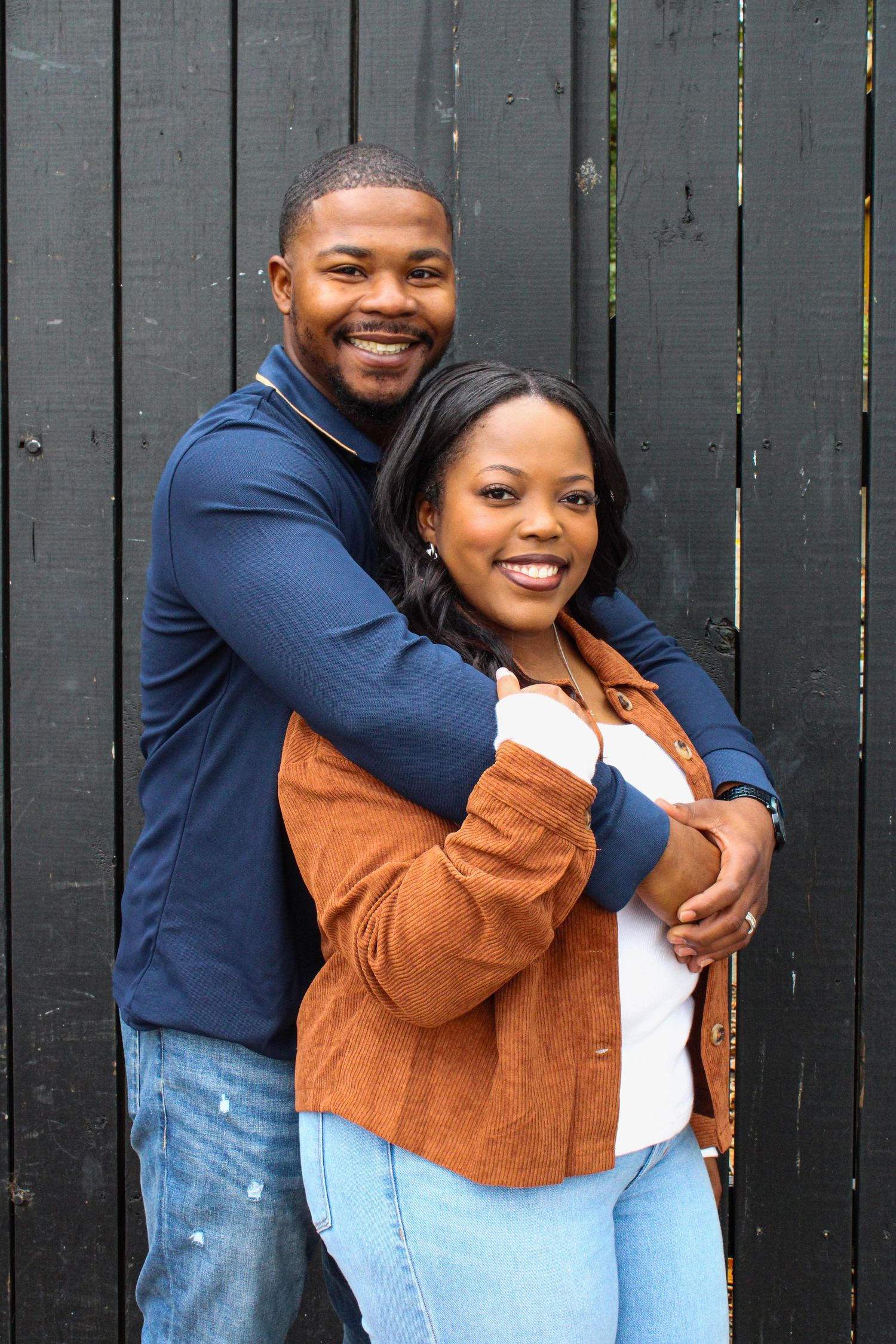 A romantic embrace against a dark fence shows a couple in coordinating brown and navy outfits.