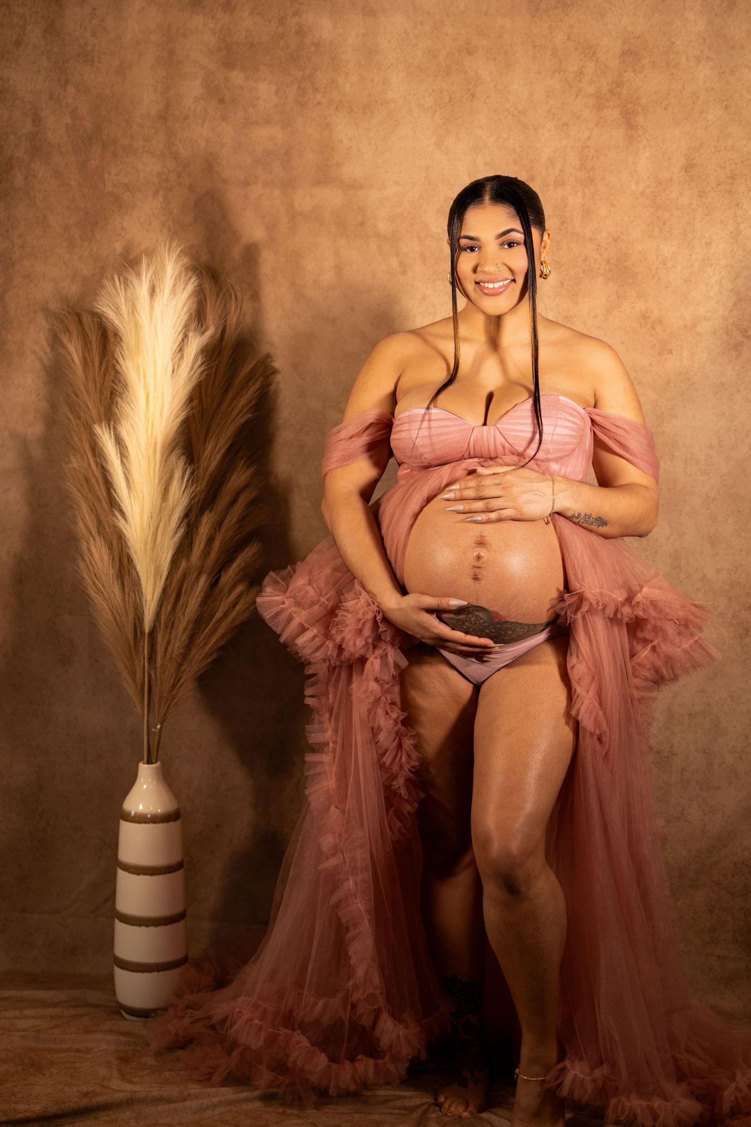 An elegant maternity boudoir photo with a dusty rose tulle robe and neutral studio backdrop.