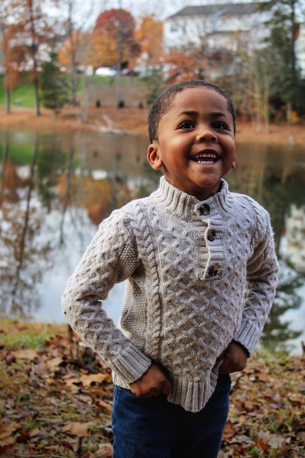 Young child wearing a cozy knit sweater smiles joyfully by a lake during fall season with autumn foliage.