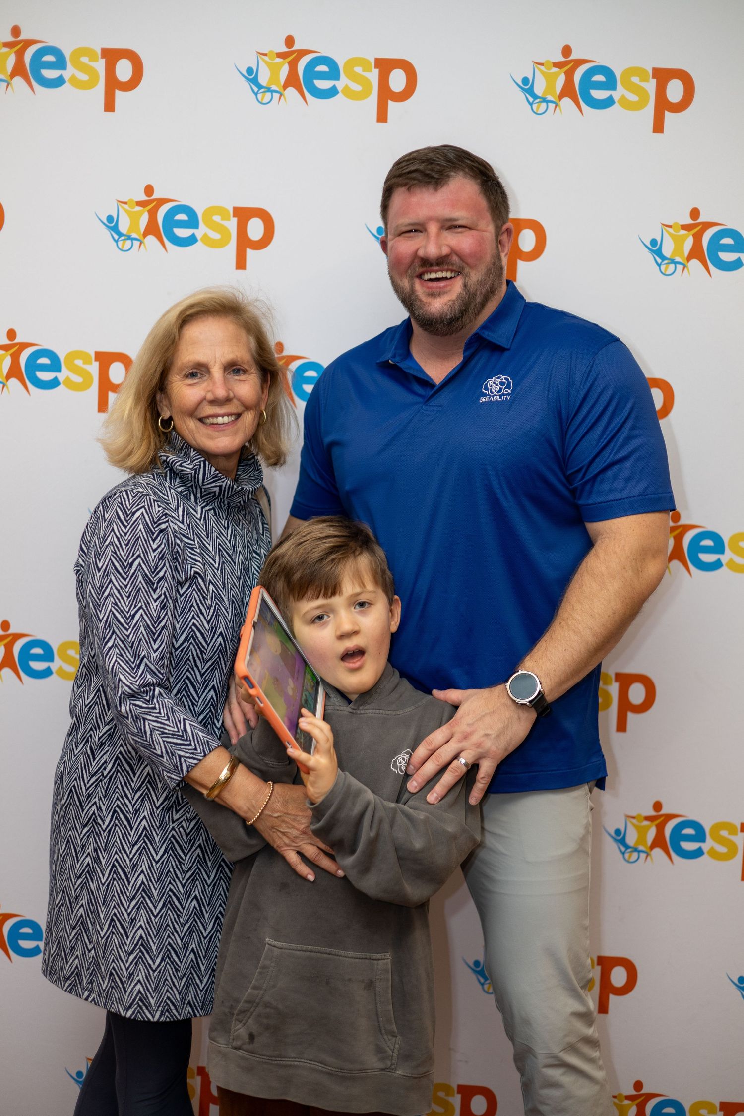 Family of three posing together at an ESP event backdrop, including a woman, young boy, and man in blue polo shirt.
