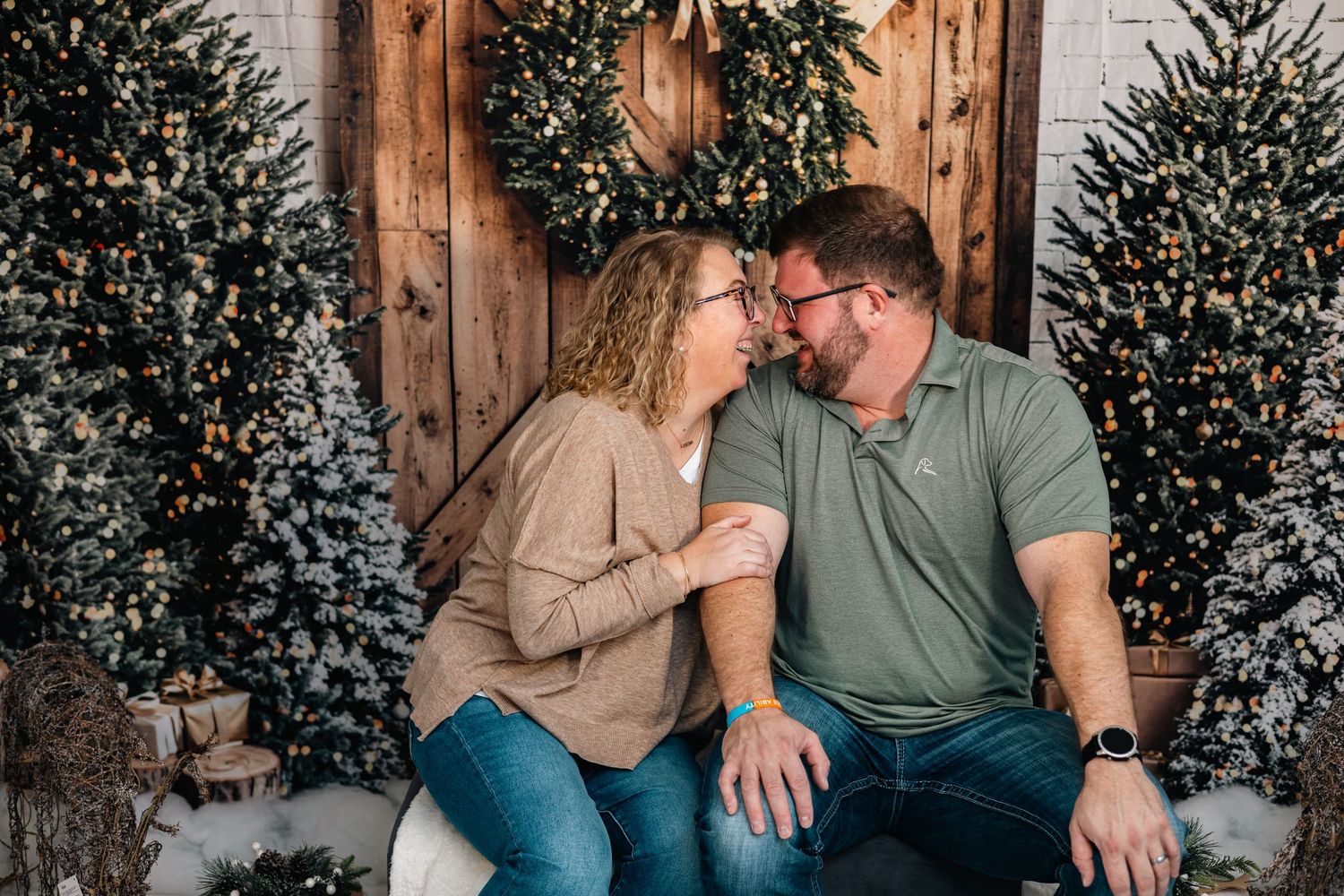 Couple sits together on rustic wooden bench with decorated Christmas trees in background during holiday photoshoot.