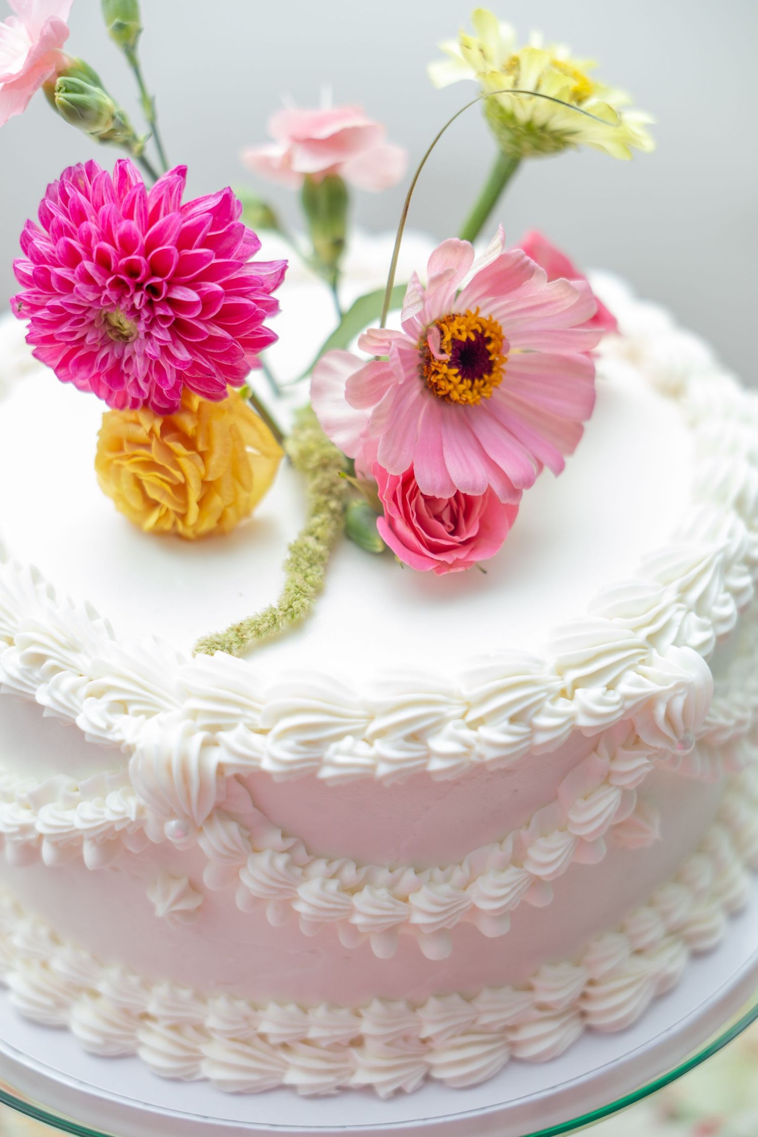 Elegant two-tier white and pink cake topped with fresh colorful flowers including dahlia, gerbera, and rose blooms.