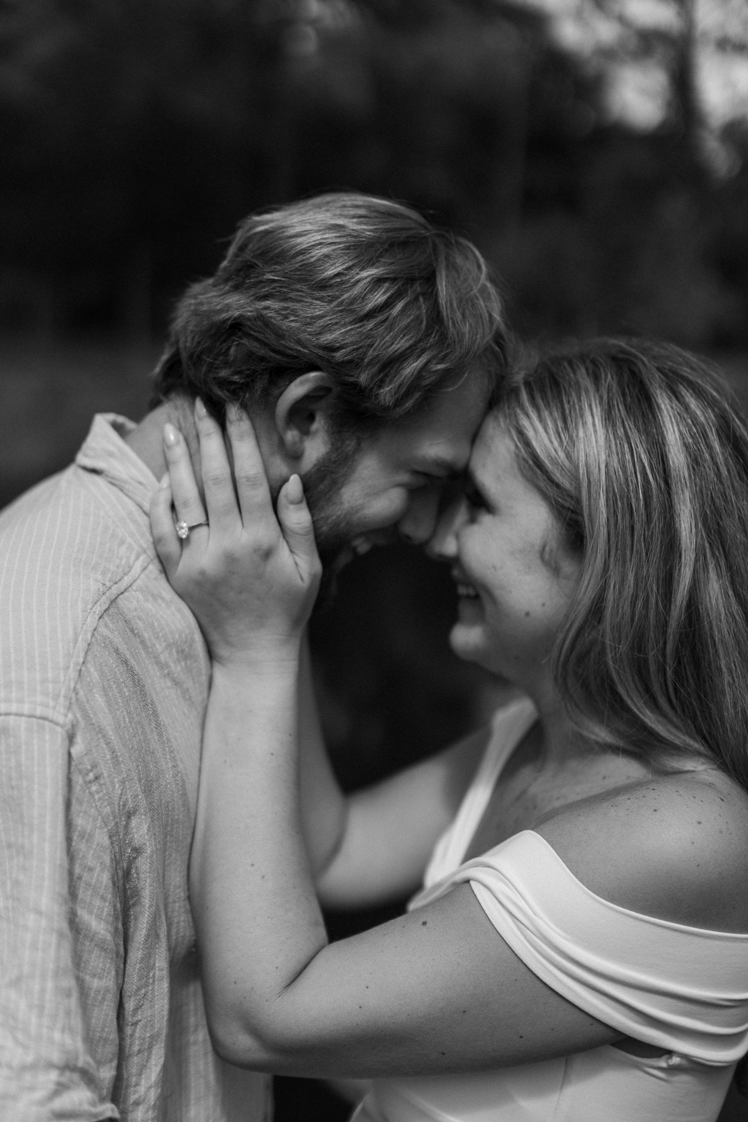 Black and white photo of a romantic couple touching foreheads, woman with engagement ring cradling man's face outdoors.