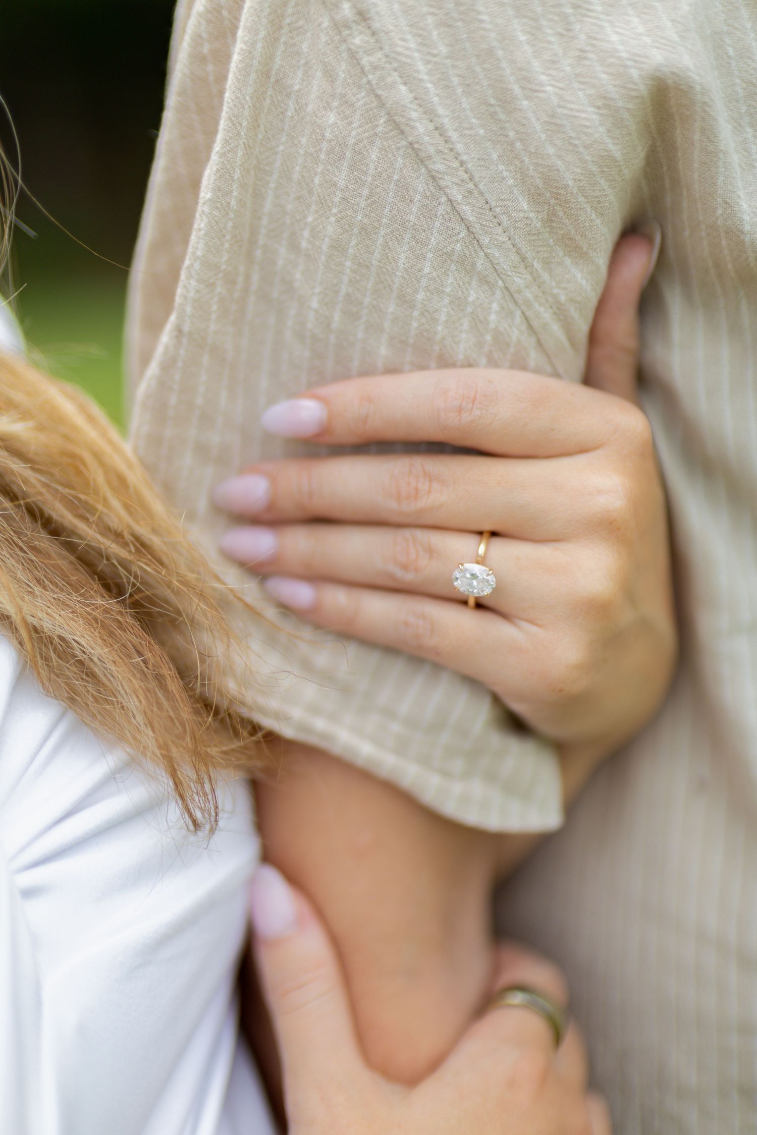 Close-up of a woman's hand wearing an oval diamond solitaire engagement ring on a gold band, resting on a man's arm.