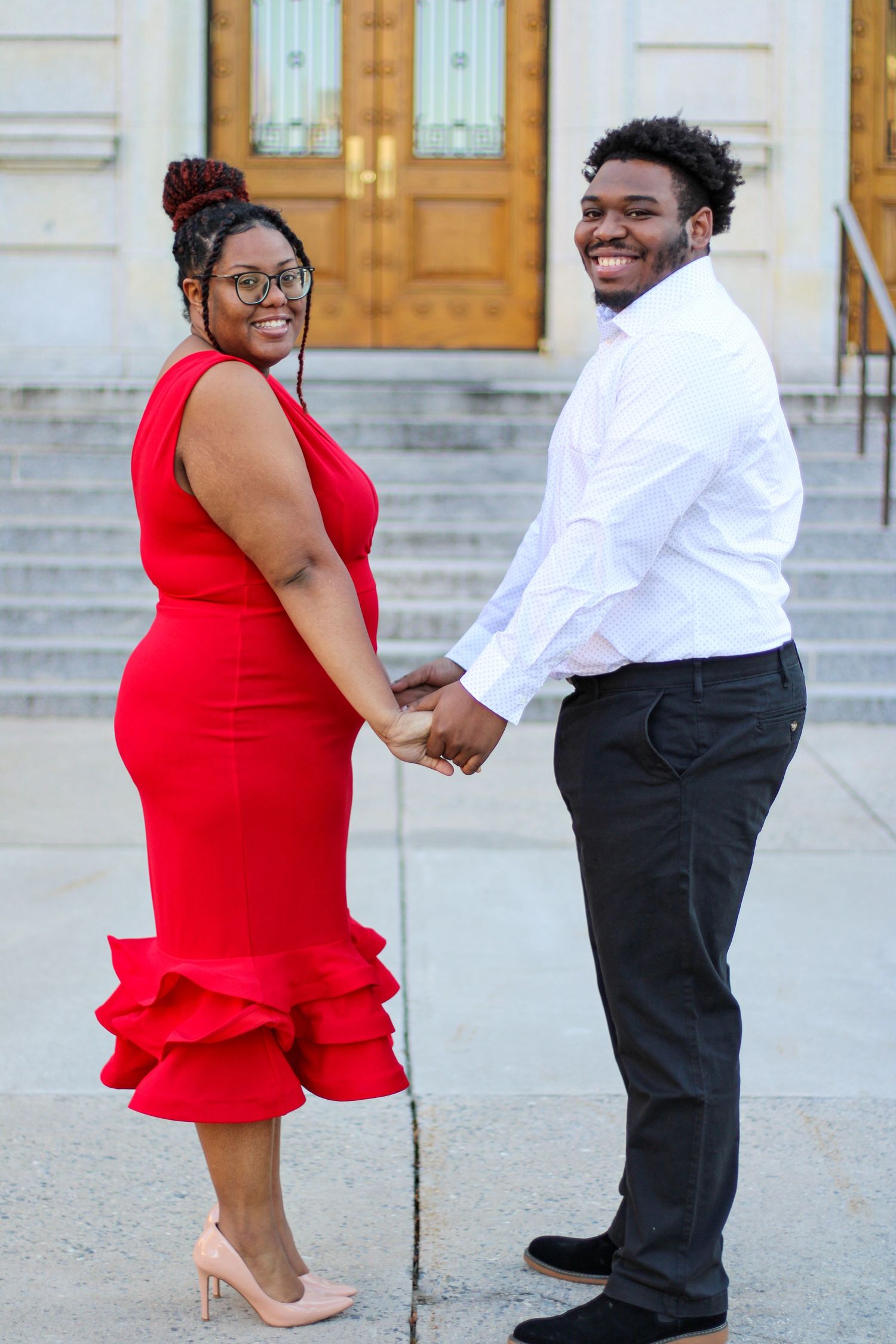 A couple in formal attire poses on stone steps, with one in a red dress and other in white shirt.