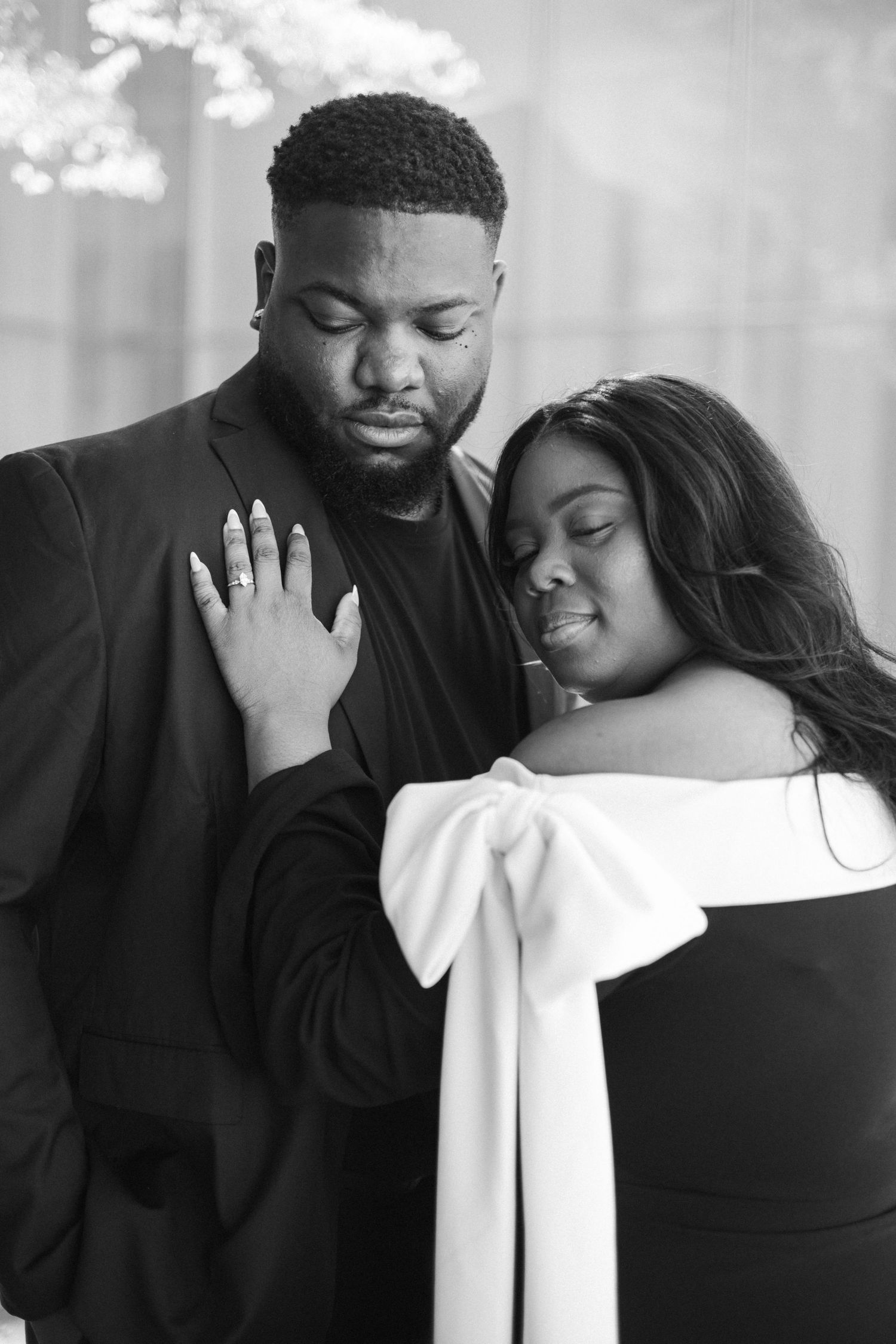 A romantic black and white portrait of a couple embracing, with the woman wearing a white bow detail dress.