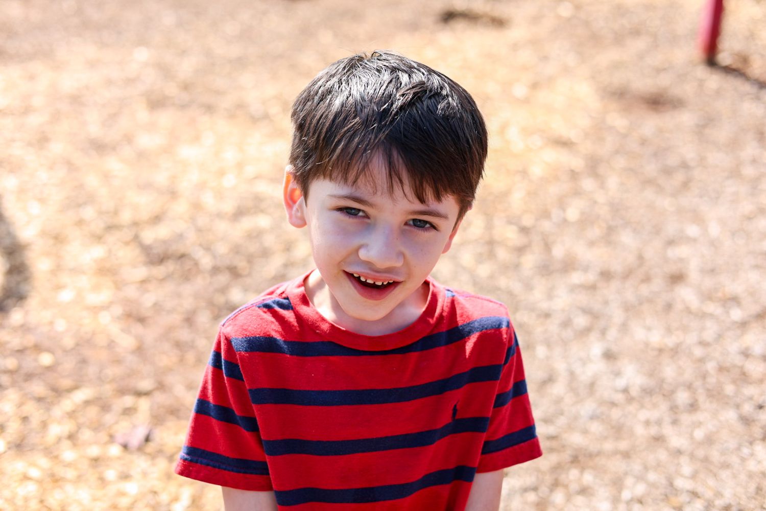 Young child in a red and navy striped t-shirt smiling brightly outdoors.
