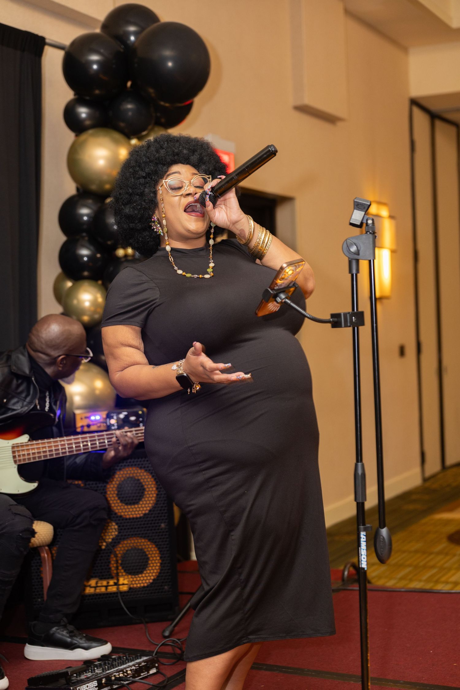A performer in a black dress sings passionately on stage with black and gold balloons decorating the background.