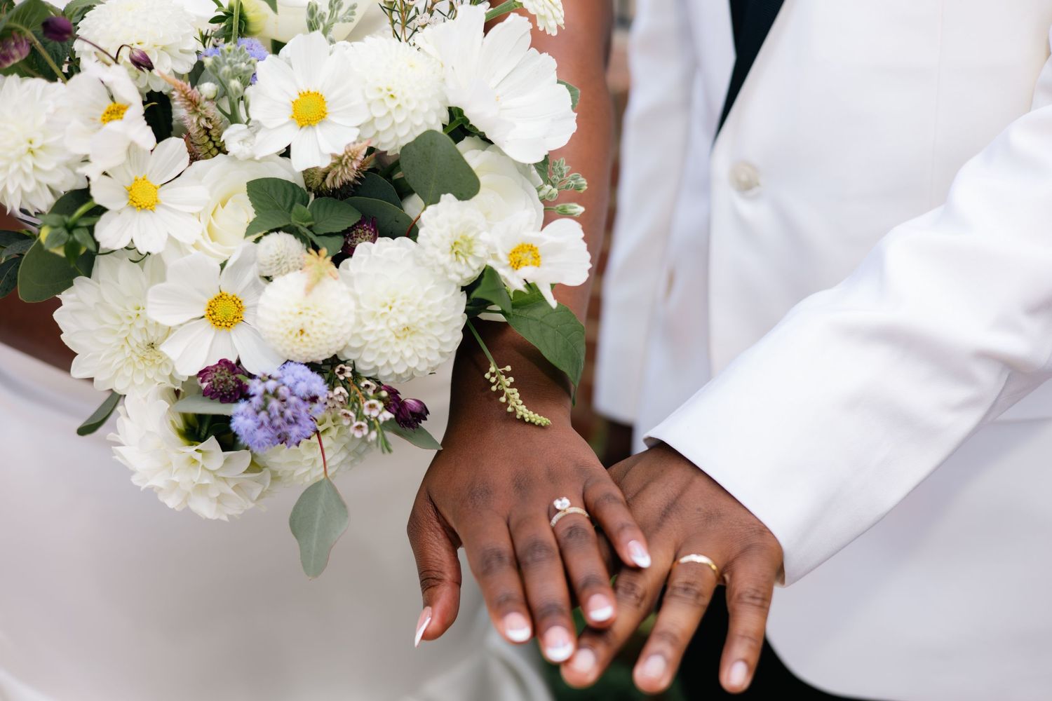 Close-up details of white bridal bouquet with daisies and purple wildflowers against white formal attire.