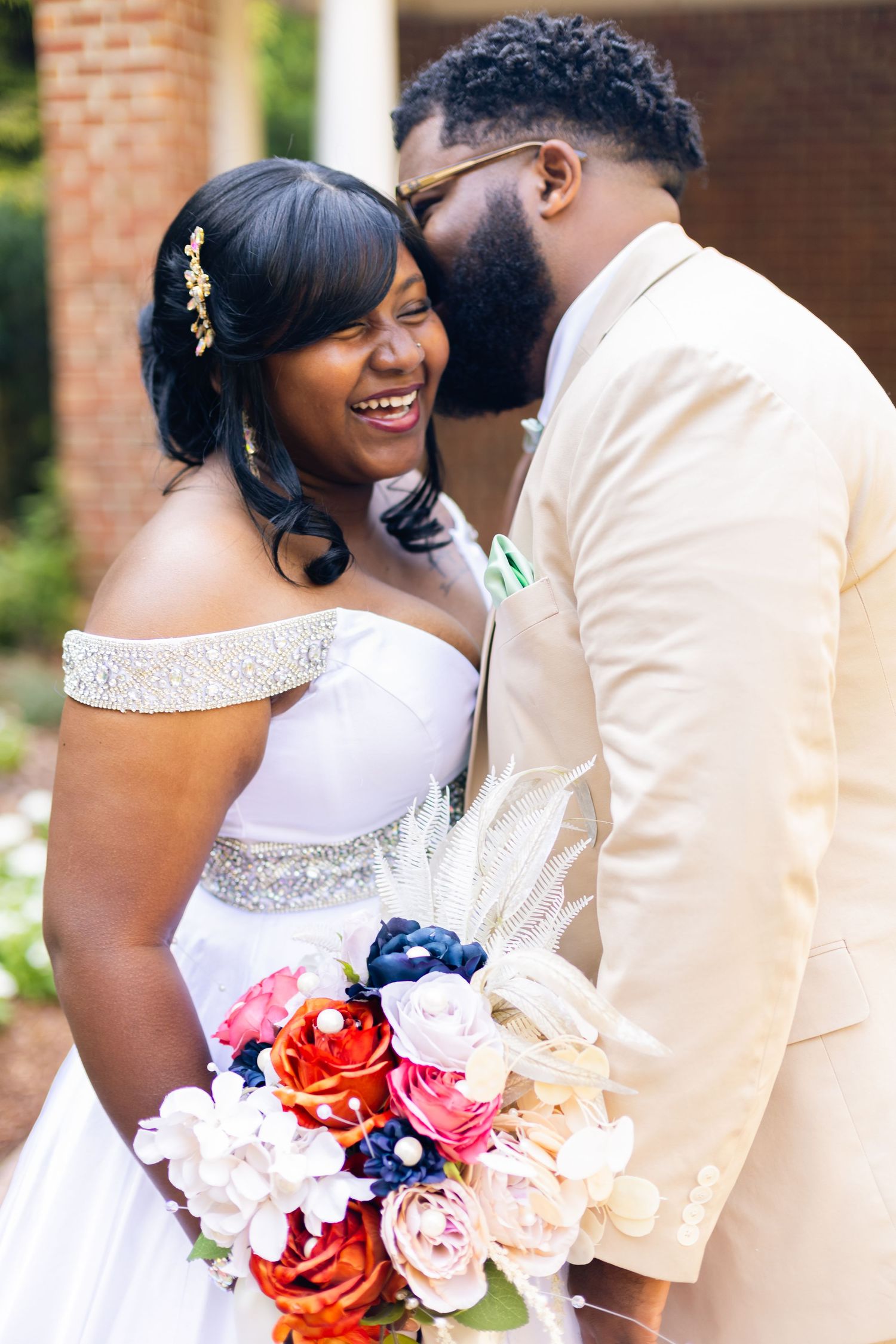 Joyful Black bride in white off-shoulder gown holds colorful bouquet as groom in beige suit whispers in her ear.