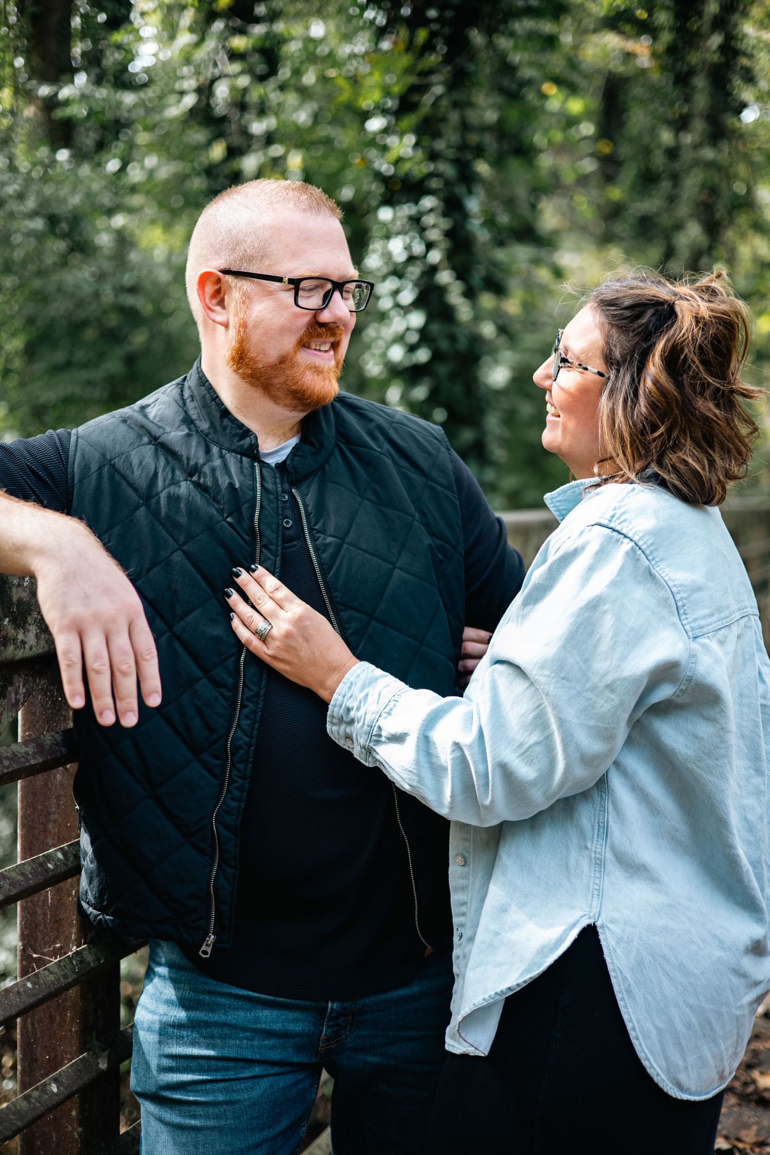 A couple in casual attire shares a tender moment on a wooden bridge surrounded by green foliage.