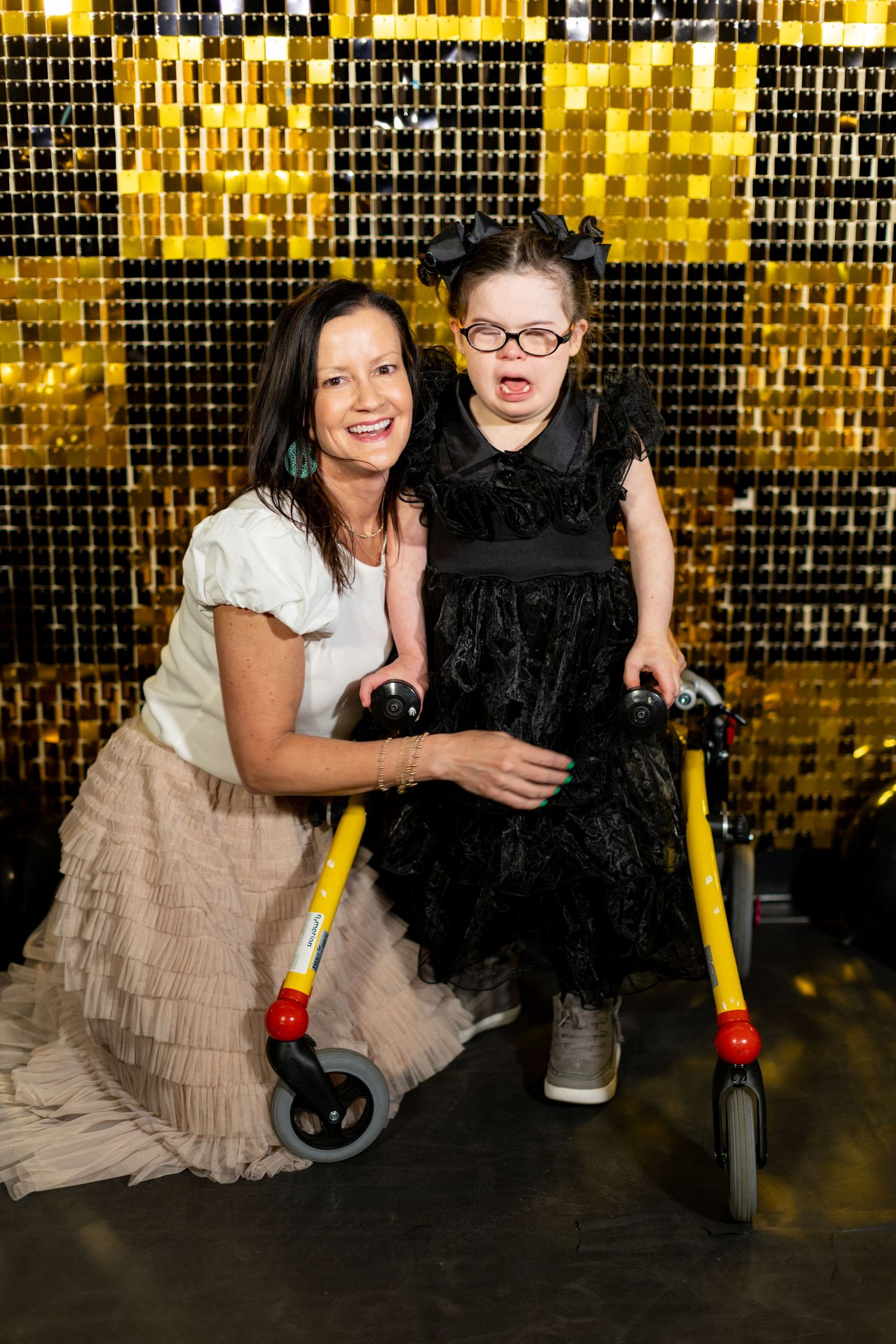 Woman and young girl with walker pose together in front of gold sequin backdrop at festive event.