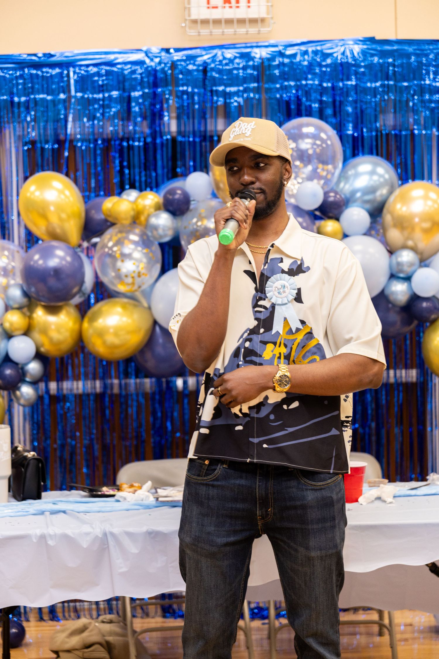 Two performers in black vests and jeans sing into microphones at an event with blue and gold balloon decorations.