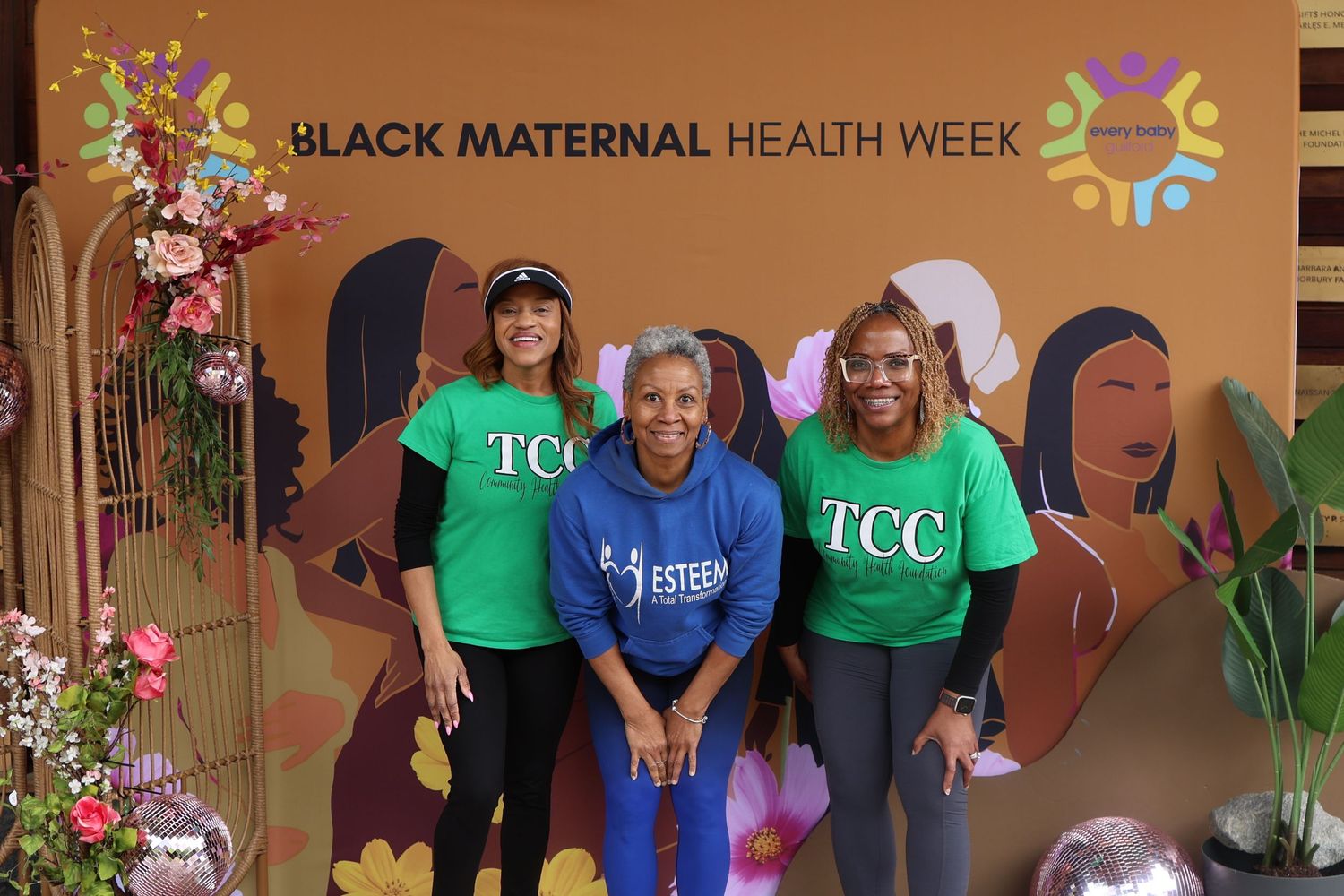 Three women in matching t-shirts pose at health awareness event with decorative backdrop.