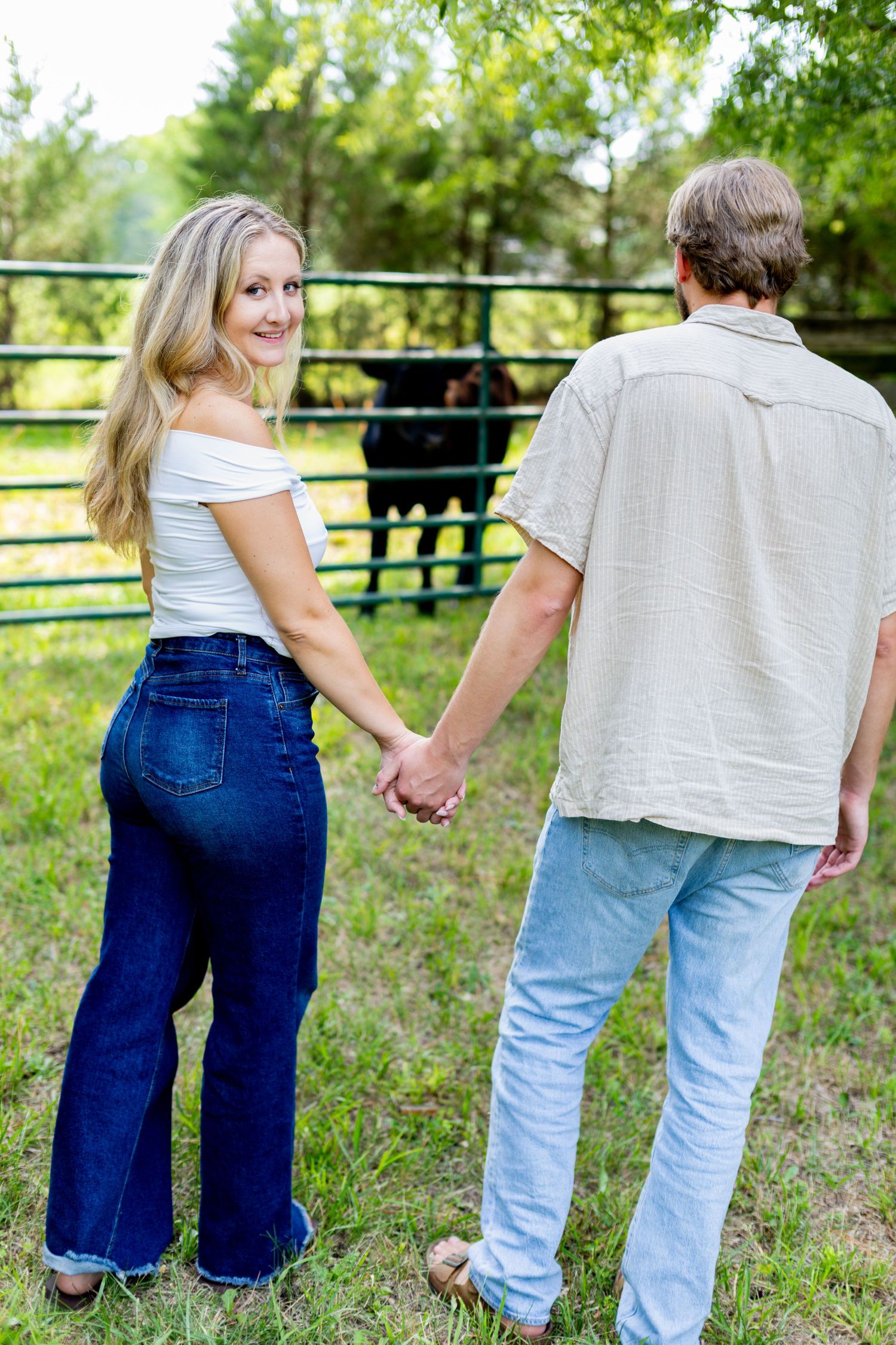 A couple holds hands while walking toward a cattle pen, with the woman looking back and smiling.