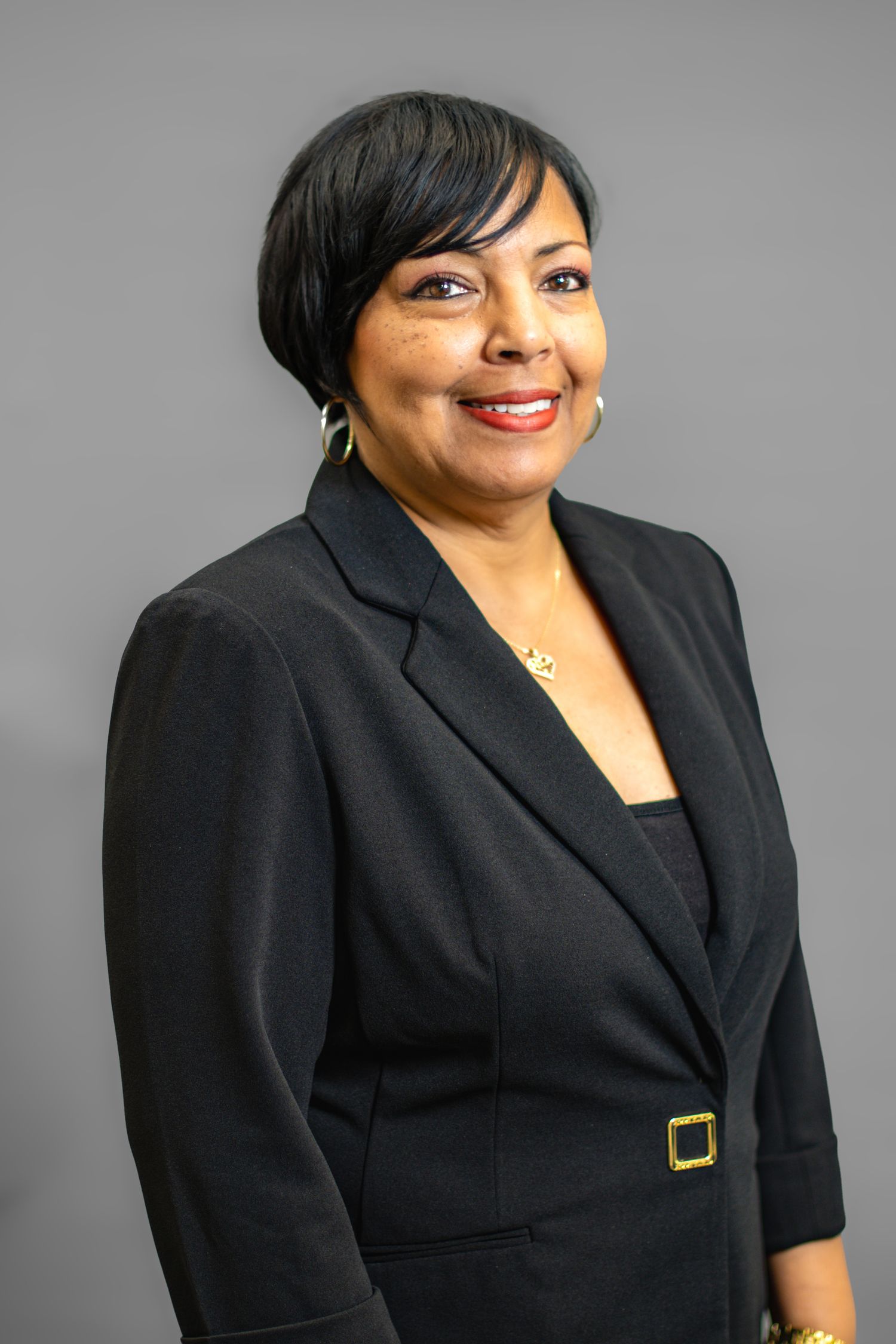 Professional woman in black blazer smiling confidently against gray background, wearing gold earrings and belt buckle.