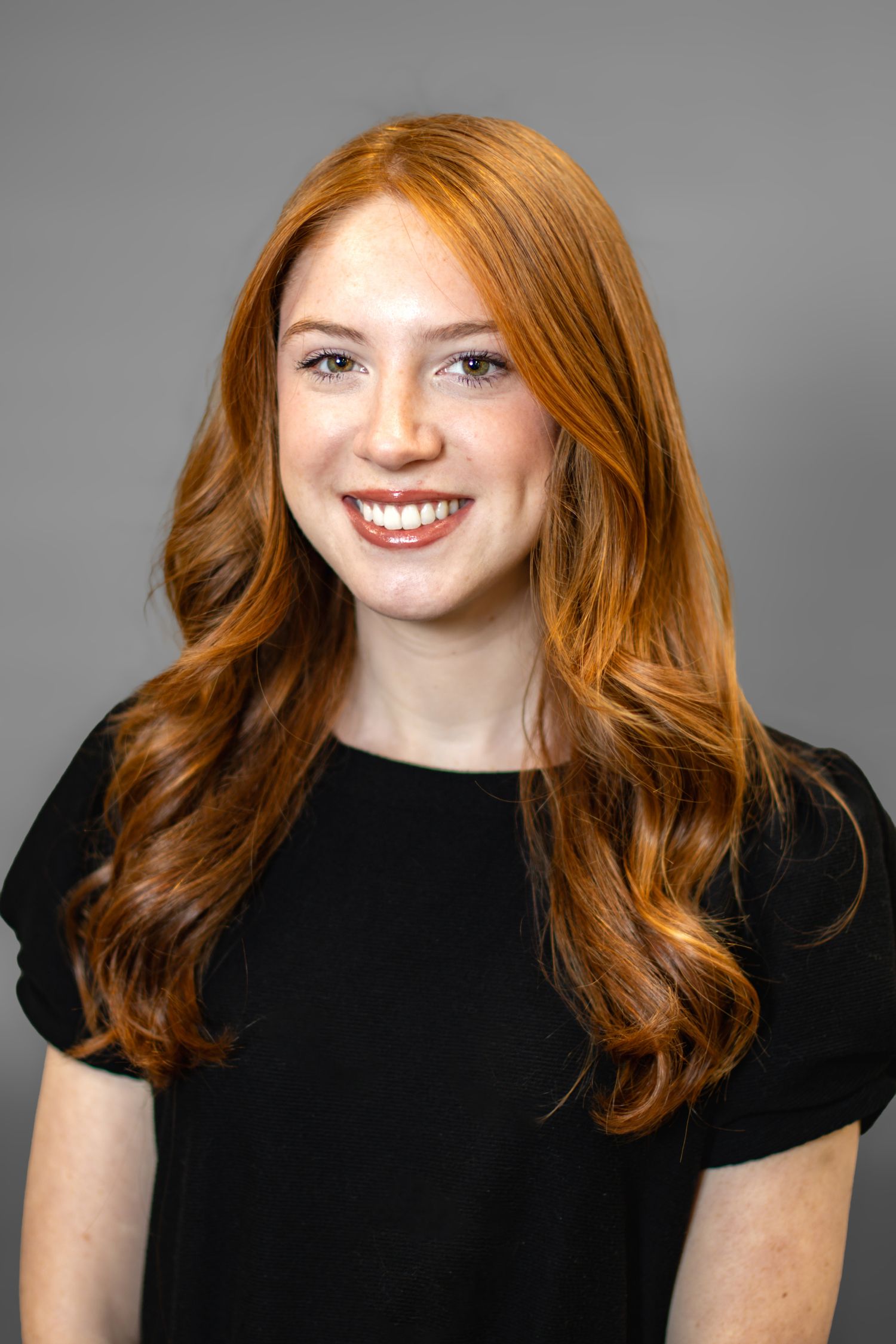 Young woman with long red hair and freckles smiling confidently in a black top against a gray background.