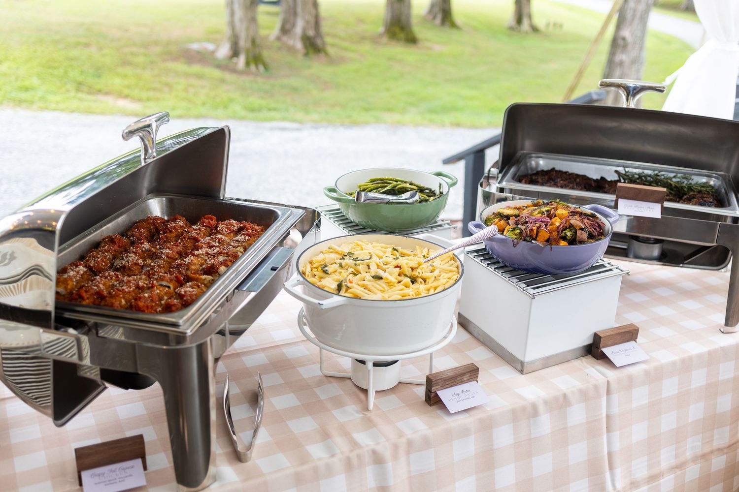 A long buffet table with stainless steel chafing dishes filled with various hot dishes at an outdoor catering event.