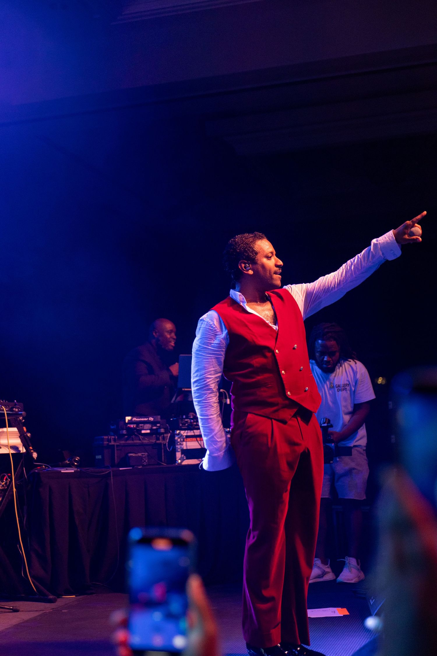 Performer in red vest and trousers points to crowd on stage under blue lighting at live concert event.