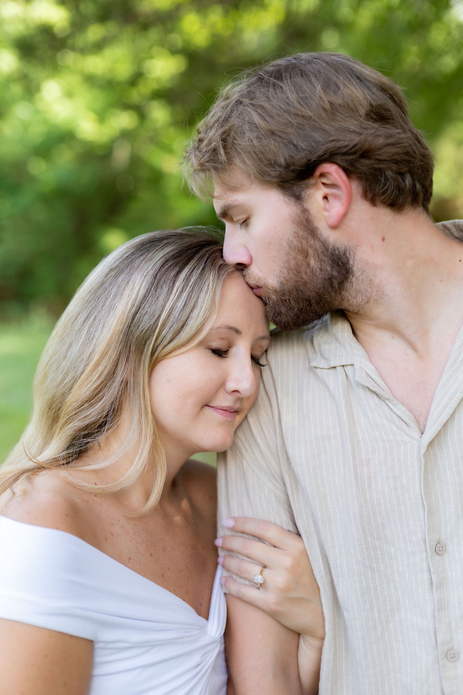 A bearded man tenderly kisses a smiling blonde woman on the head outdoors in a lush green park setting.