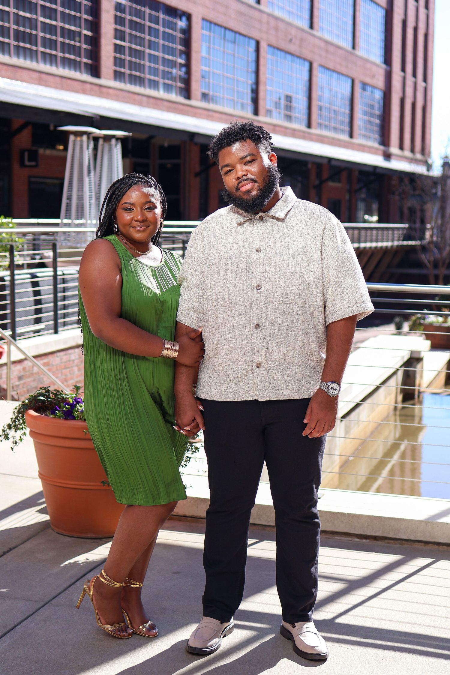 A couple poses together on an outdoor deck with potted plants and industrial architecture.