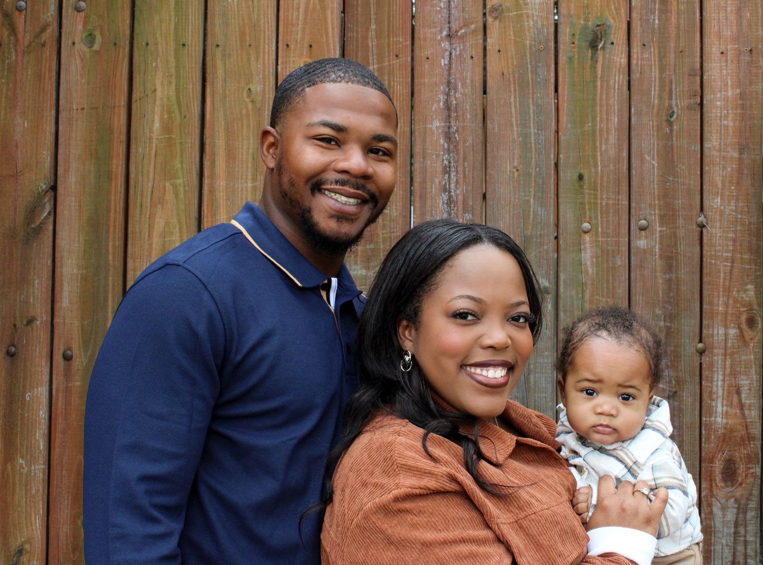 Family of three poses together in front of wooden fence for portrait.