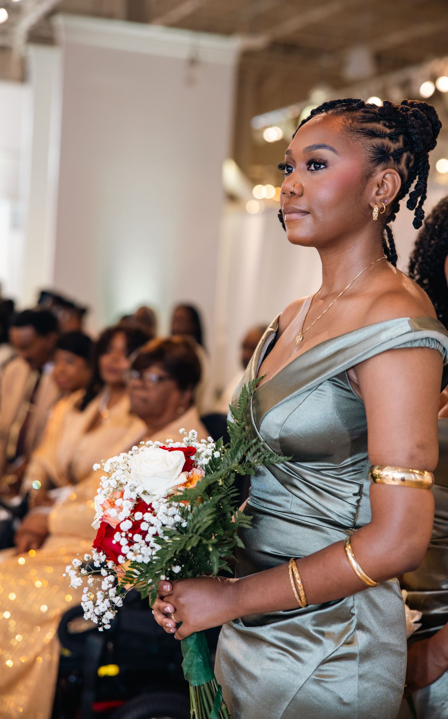 Bridesmaids in elegant sage green satin dresses hold bouquets during an indoor wedding ceremony.