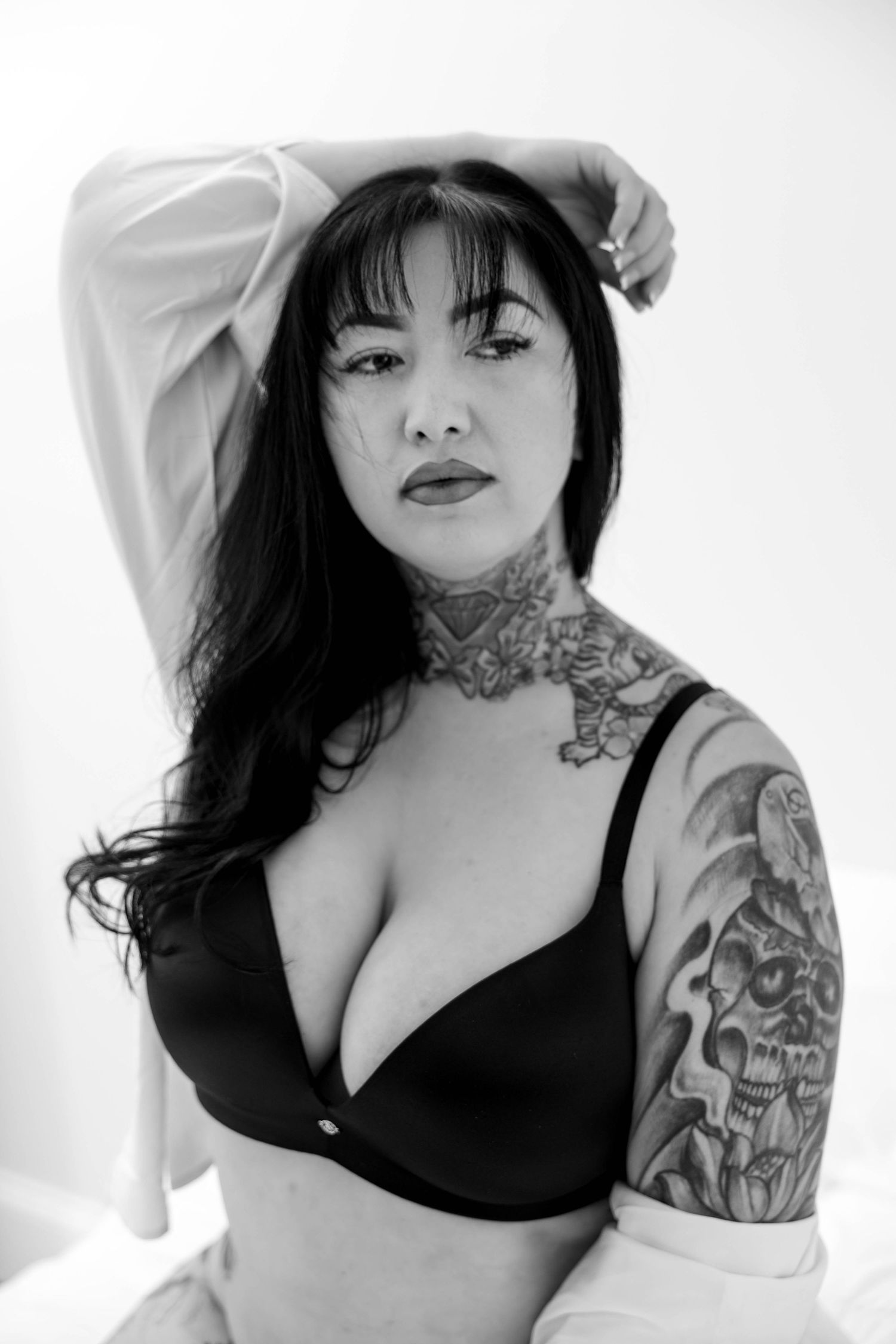 Black and white portrait of a model in black bra with visible neck and arm tattoos against white background.